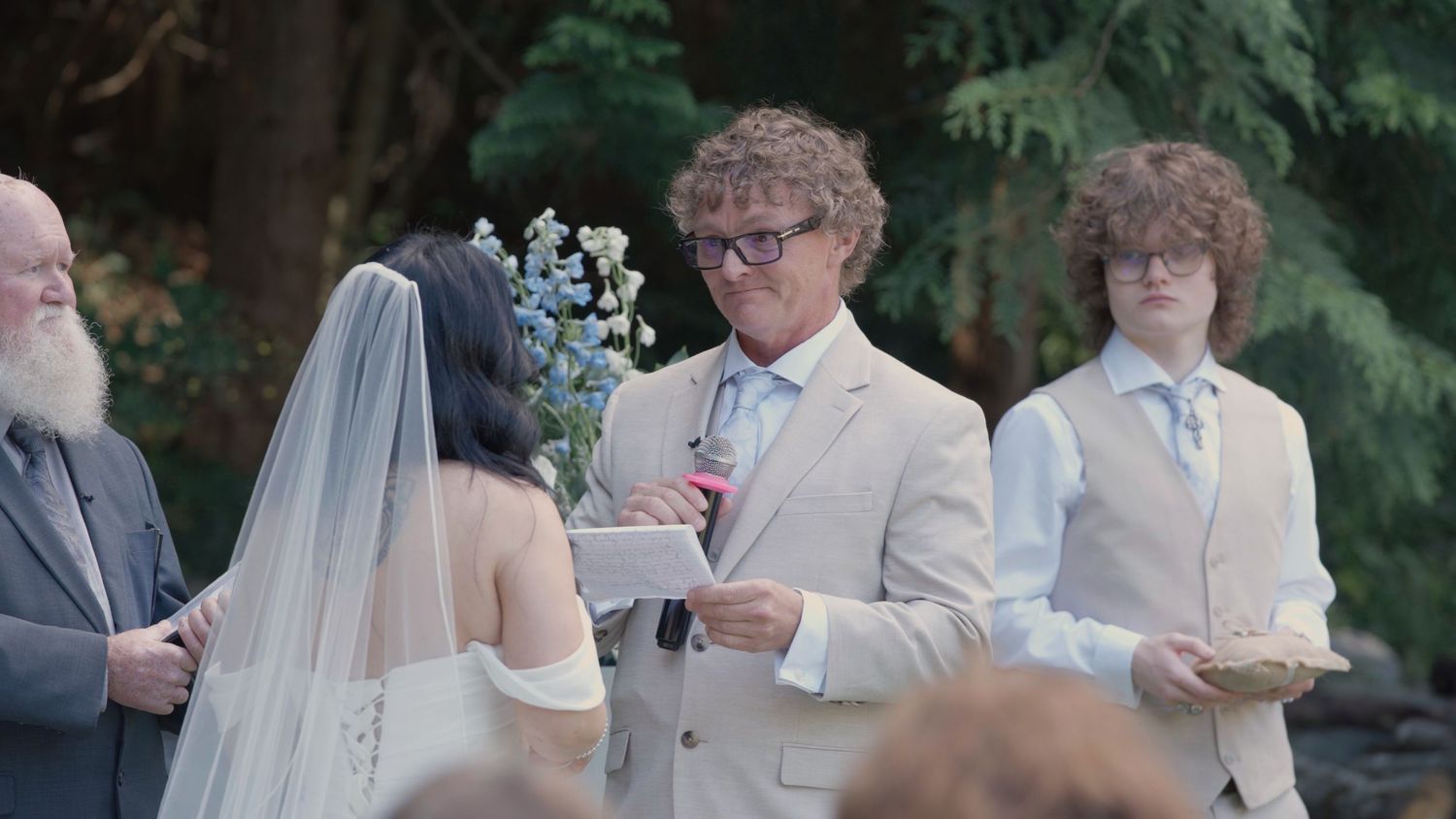 Groom reads vows to bride at outdoor wedding ceremony surrounded by guests and officiant.