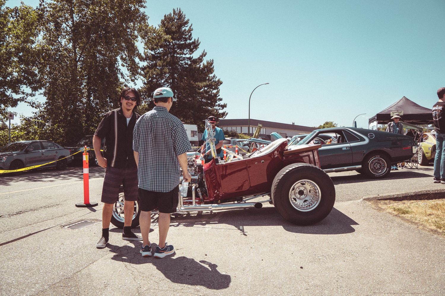 Two people stand beside a vintage drag racing car with open hood at an outdoor car show event.