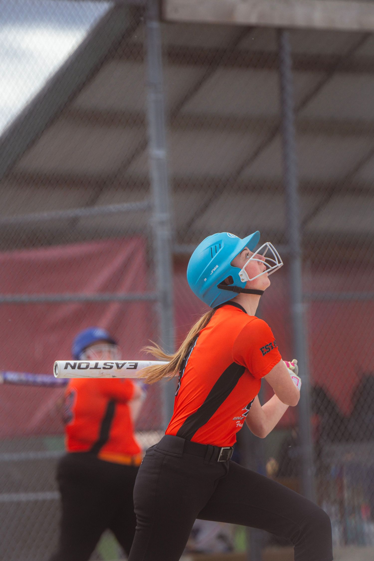 Softball player in orange jersey and teal helmet running after hitting with Easton bat.