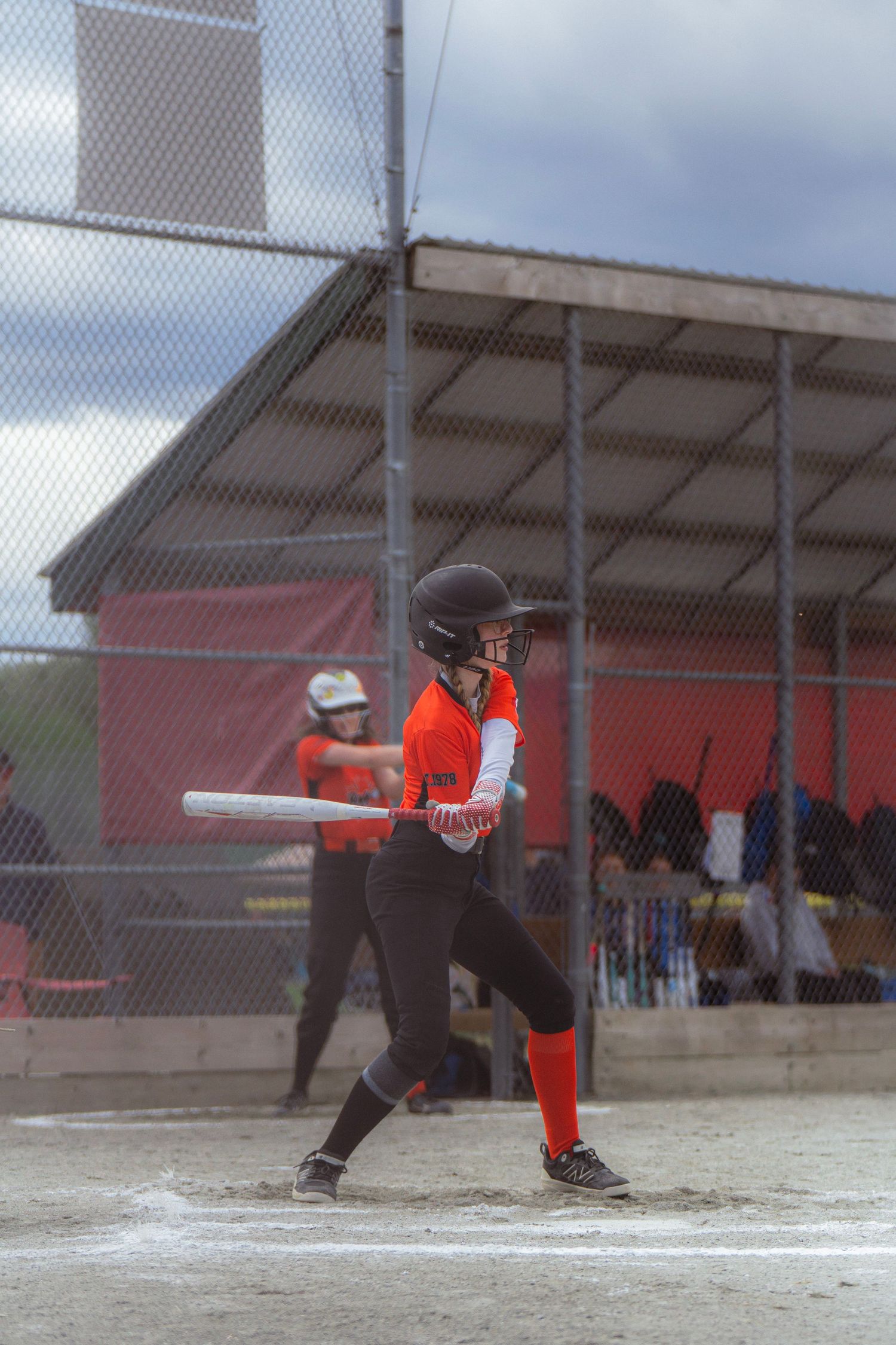 Young softball batter in orange uniform swings at a pitch during a game at an outdoor diamond.