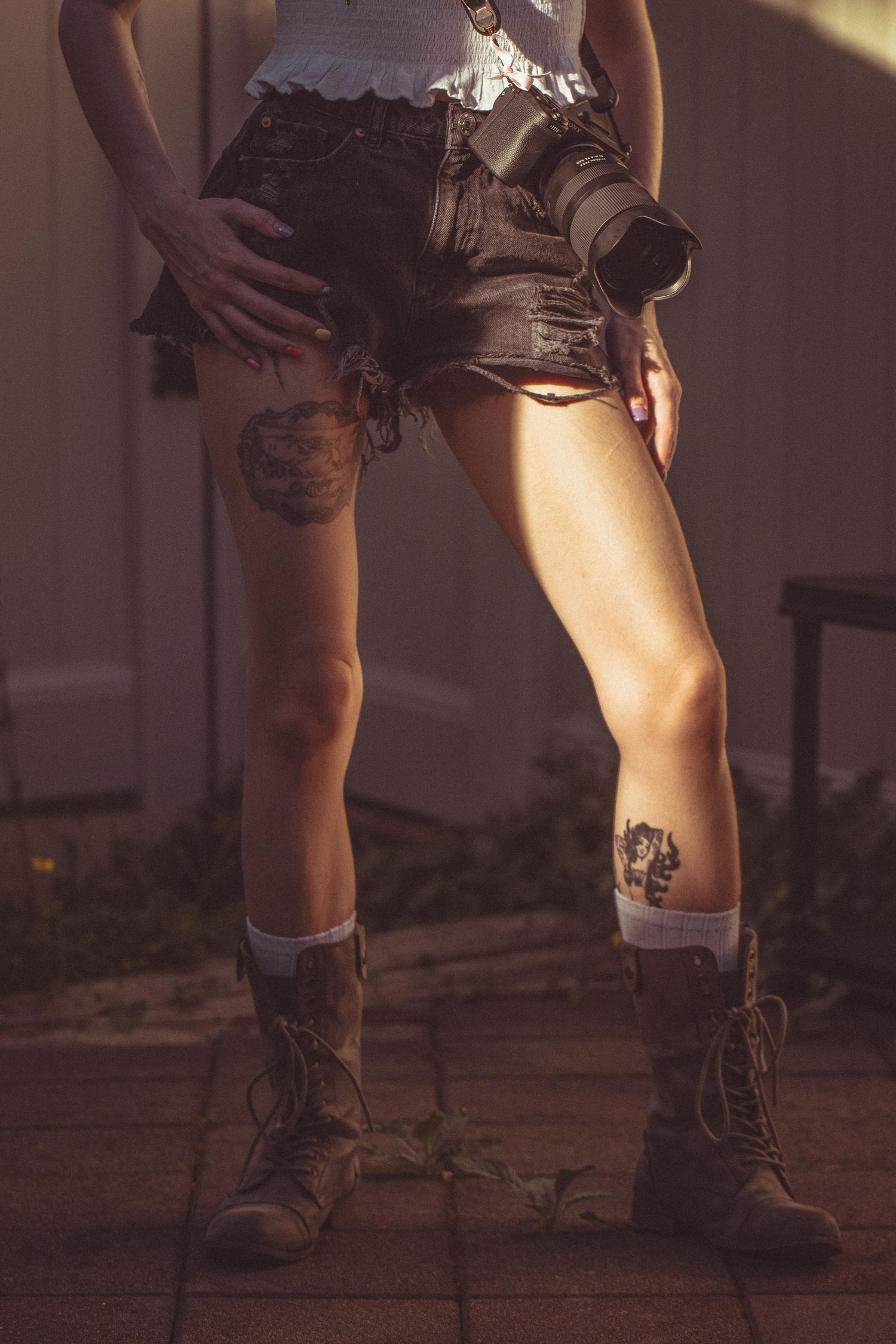 Close-up of tattooed legs of woman wearing denim shorts and brown combat boots holding a DSLR camera outdoors.