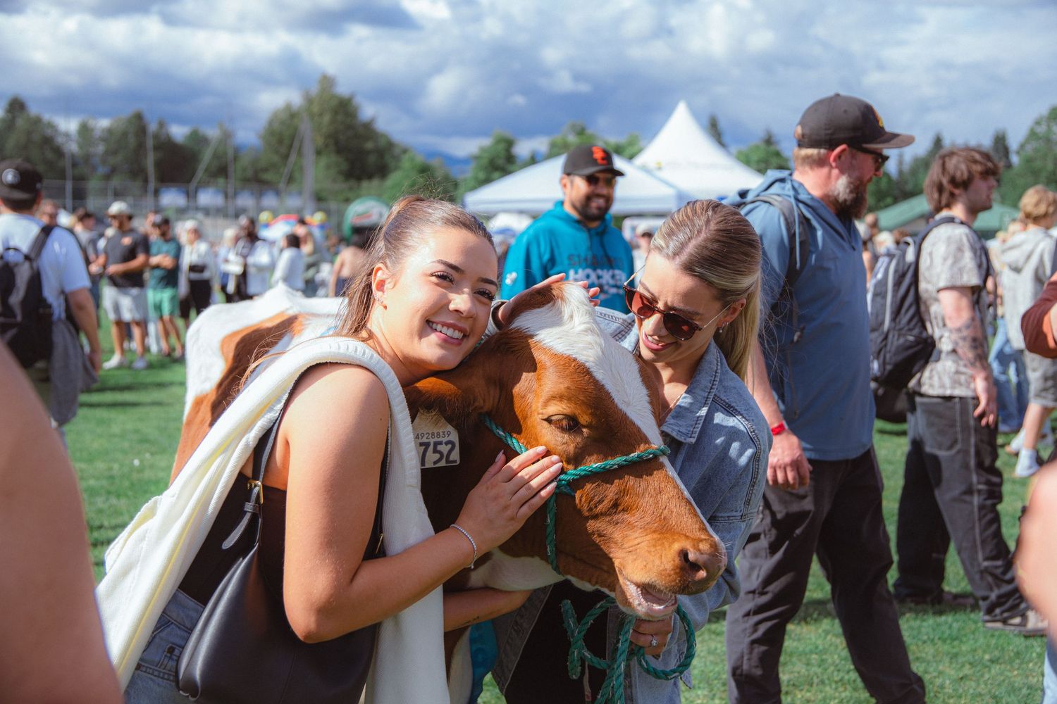 Two smiling women pose with a brown dog at a sunny outdoor festival event.