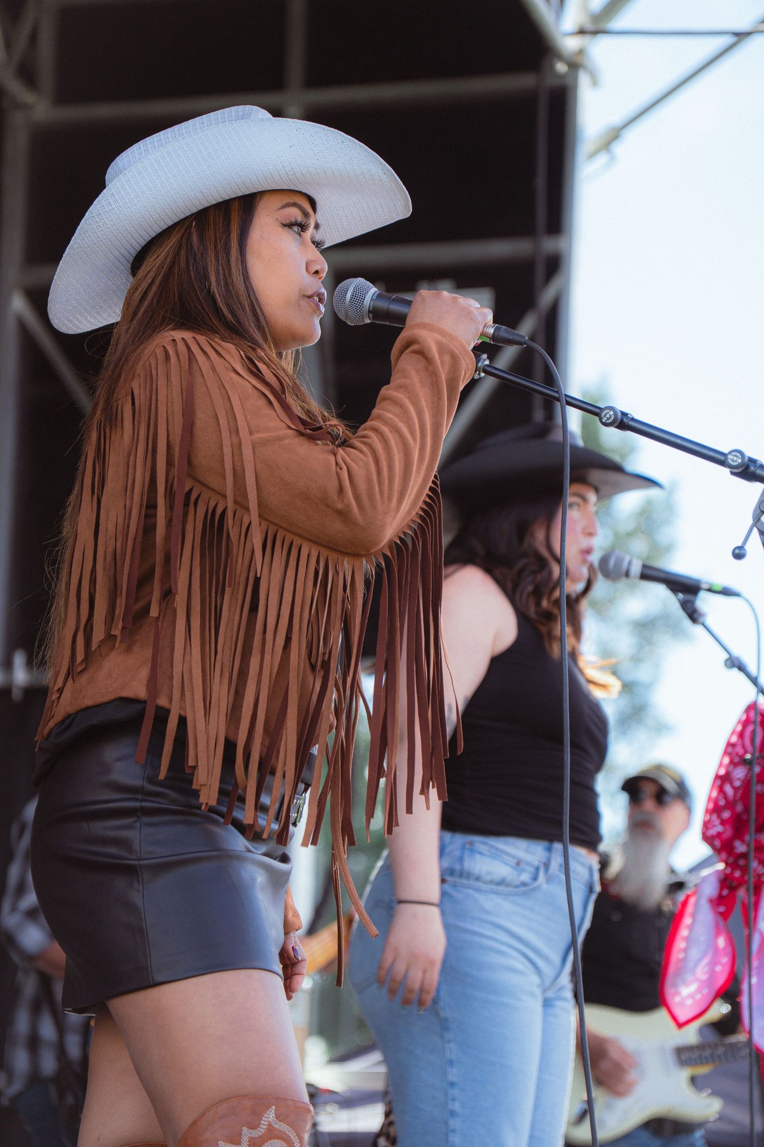 Female vocalist in white cowboy hat and brown fringe jacket sings at outdoor country music festival.