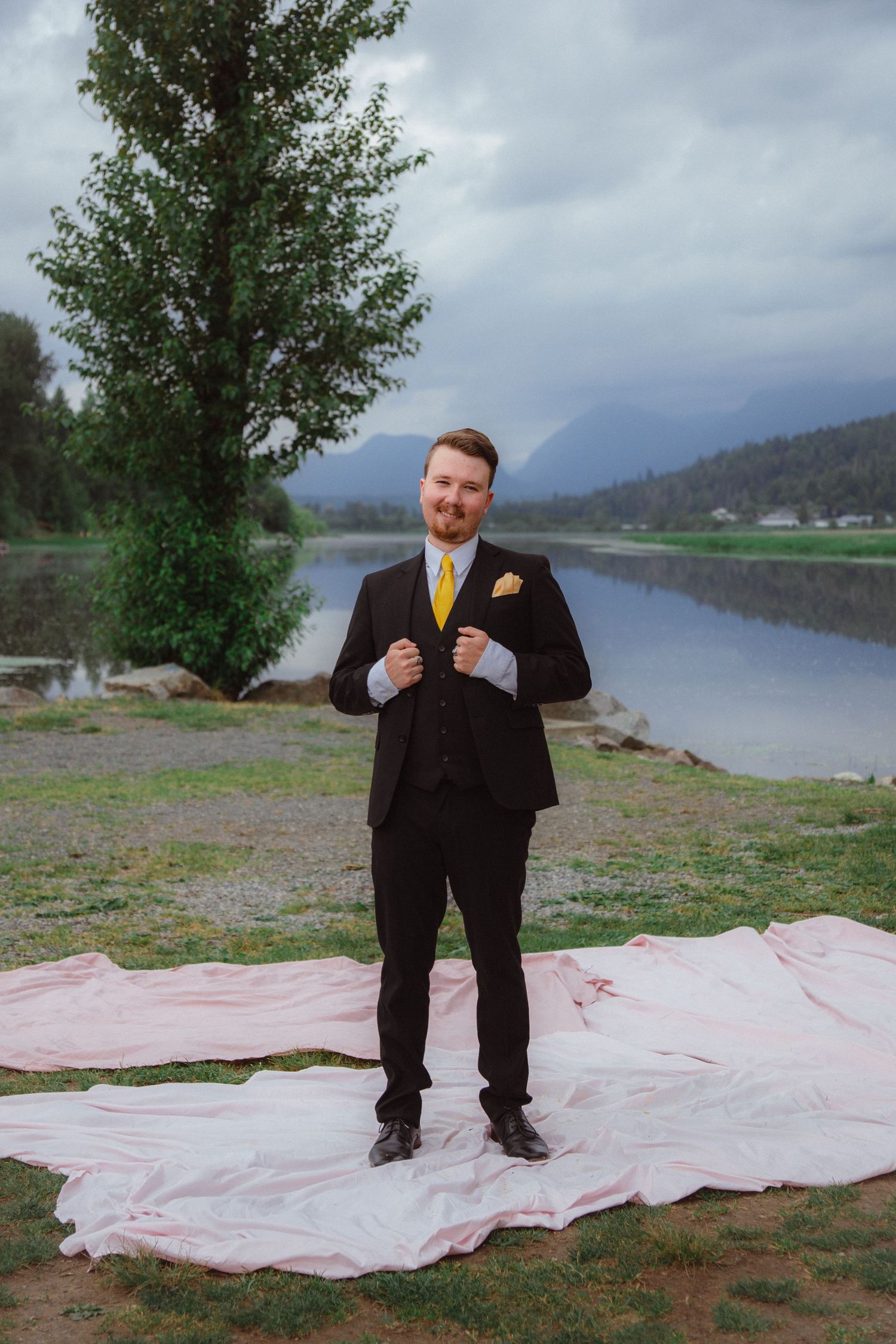 Groom in black suit with yellow tie stands on pink fabric by a mountain lake.