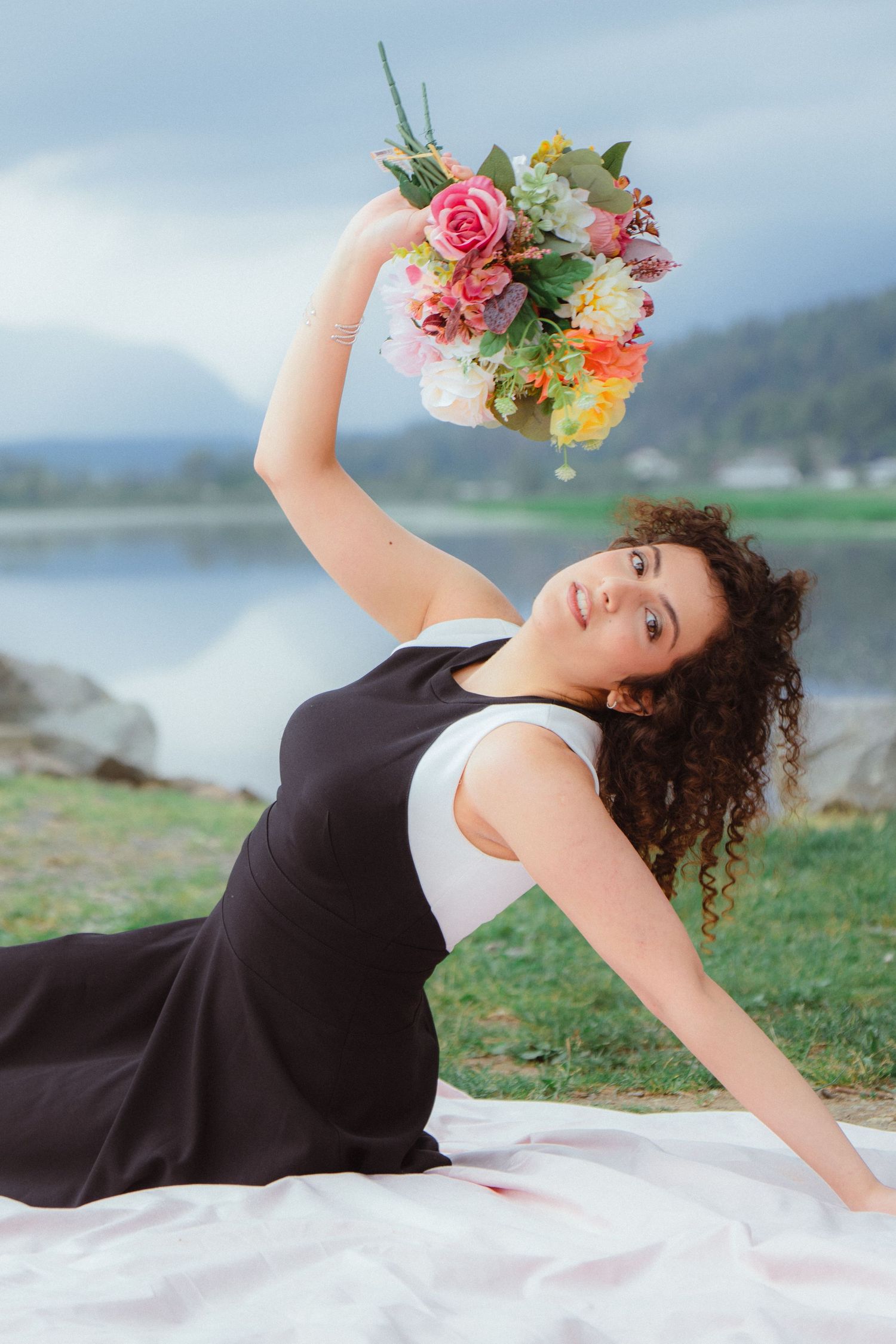 Curly-haired woman in black dress posing on blanket holding colorful bouquet aloft by mountain lake.