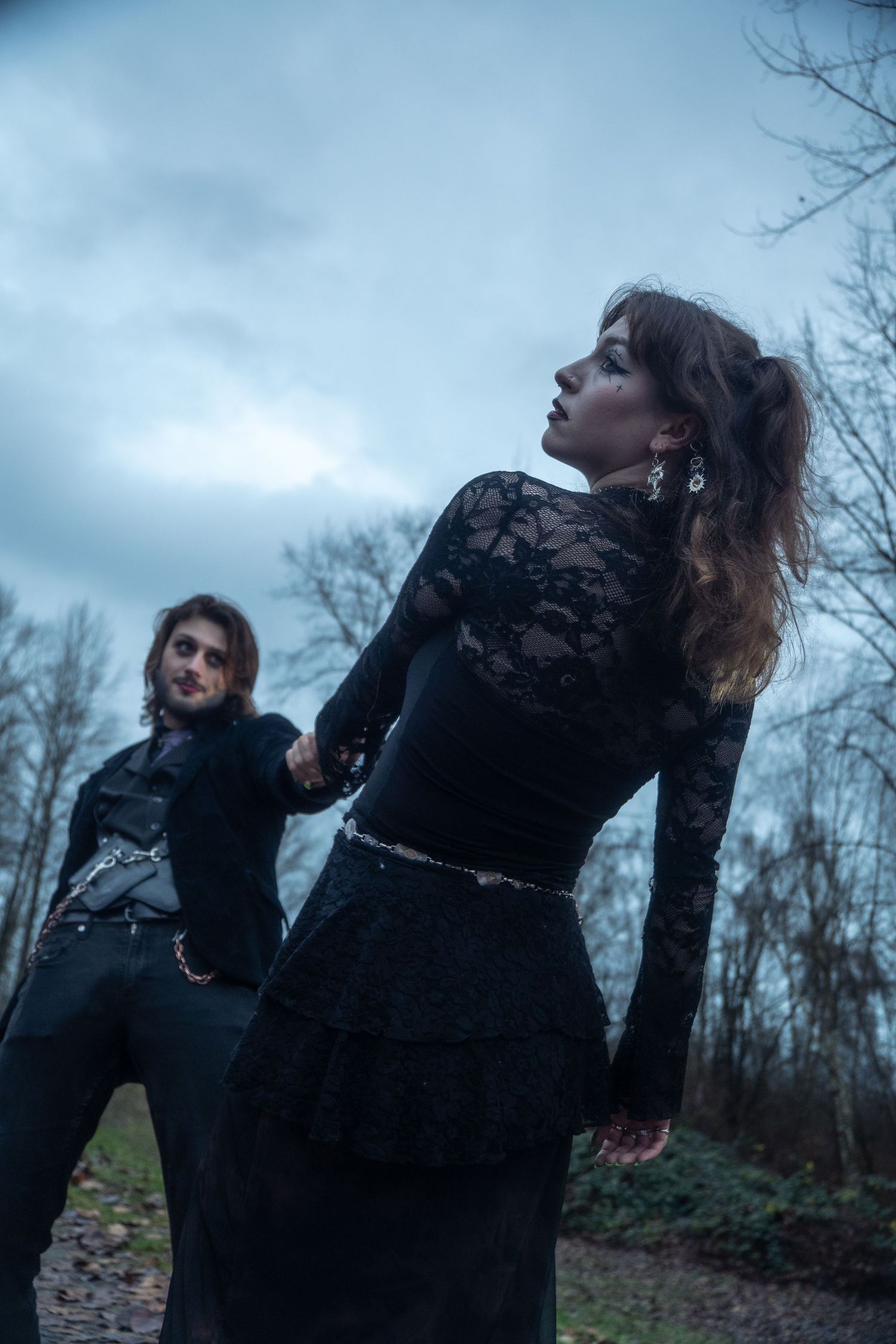 Gothic couple in black hold hands on a forest path under dramatic cloudy sky.