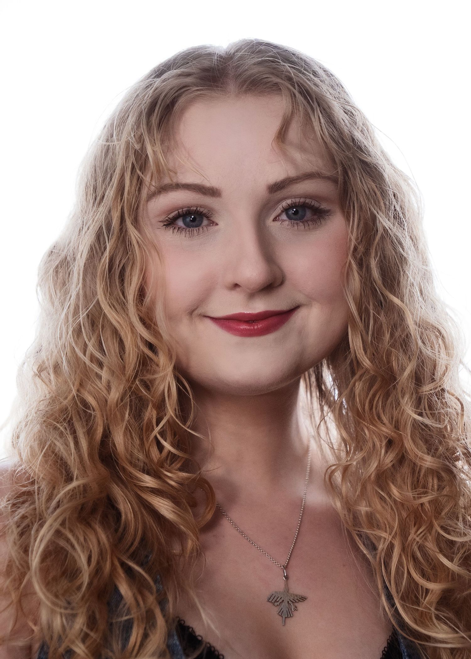 Smiling young woman with long curly blonde hair and red lips wearing a necklace, professional headshot.