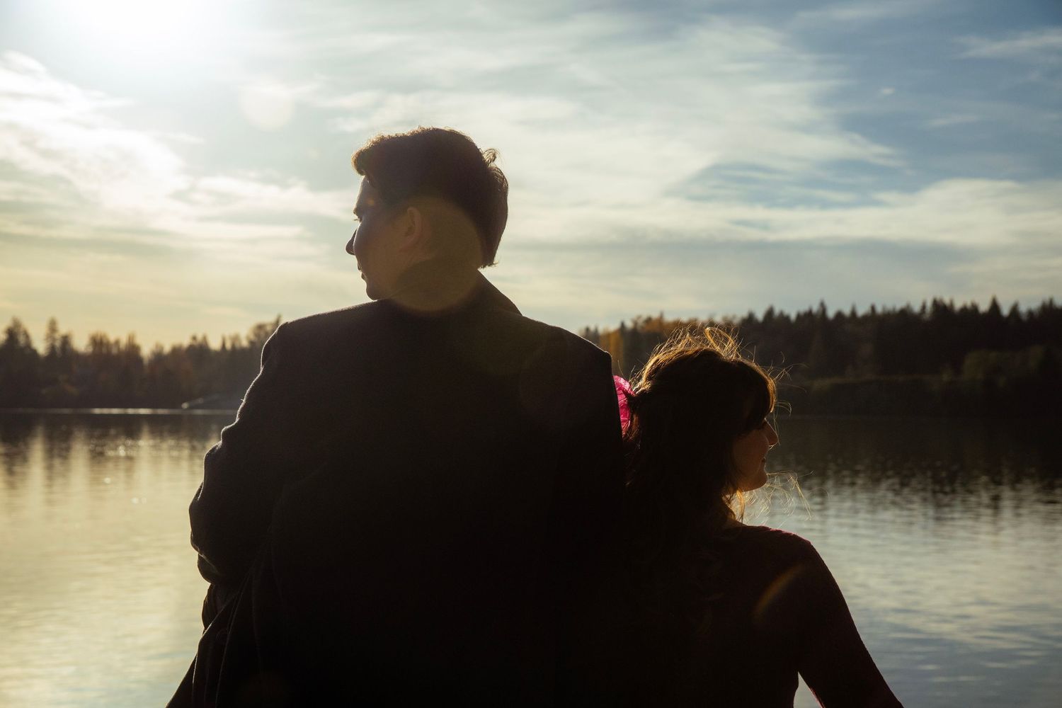 Silhouetted couple sitting together gazing at a tranquil lake under a dramatic cloudy sky at sunset.