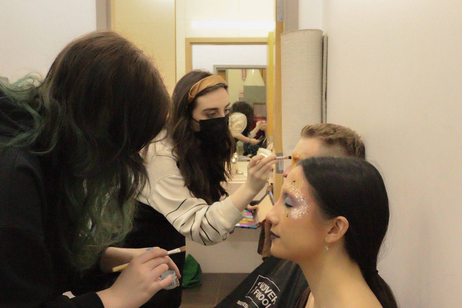 Two makeup artists apply colorful theatrical makeup to a performer backstage before a show.