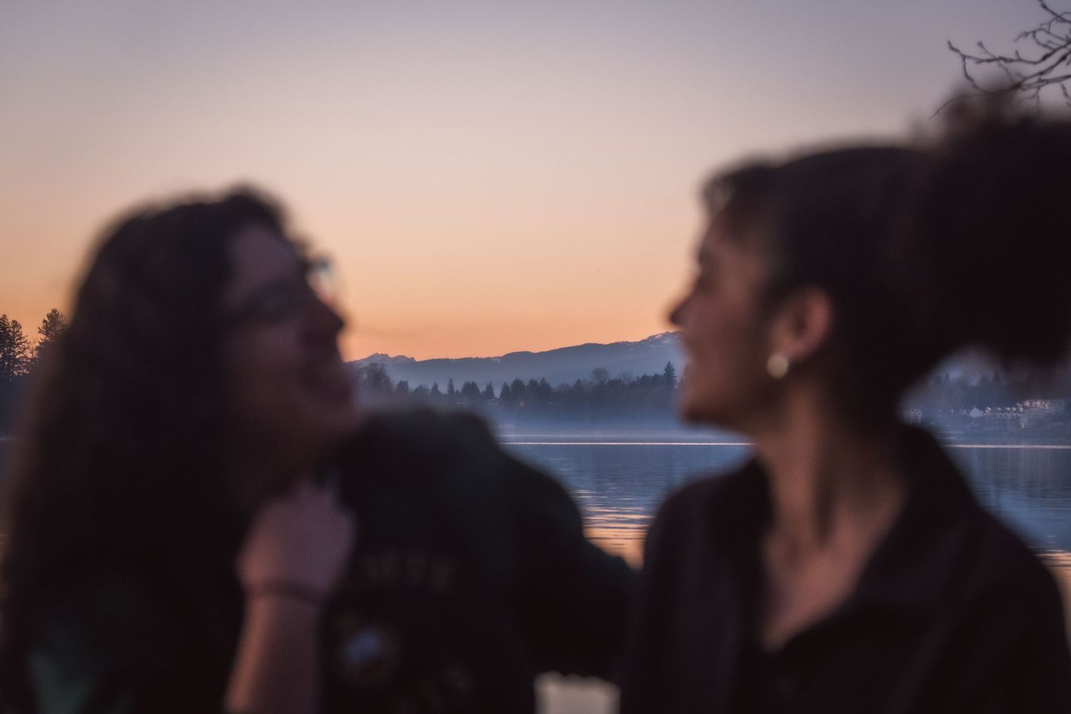 Two women share a tender moment by a tranquil lake at sunset, one gazing at the other in soft golden light.