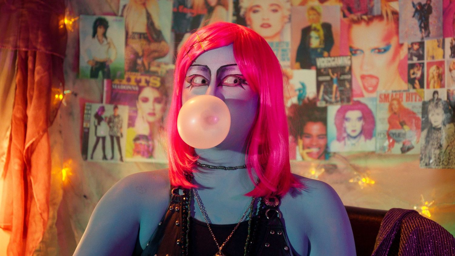 Person with pink wig and blue makeup blowing a large bubble gum bubble in front of pop culture wall posters.