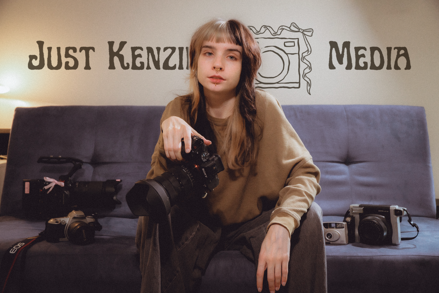 Young photographer holding a camera sits on a couch beneath a Just Kenzie Media logo sign.
