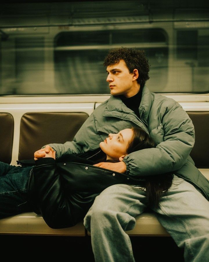 Couple sitting on subway bench, woman resting head on man's lap, both wearing green puffer jackets.