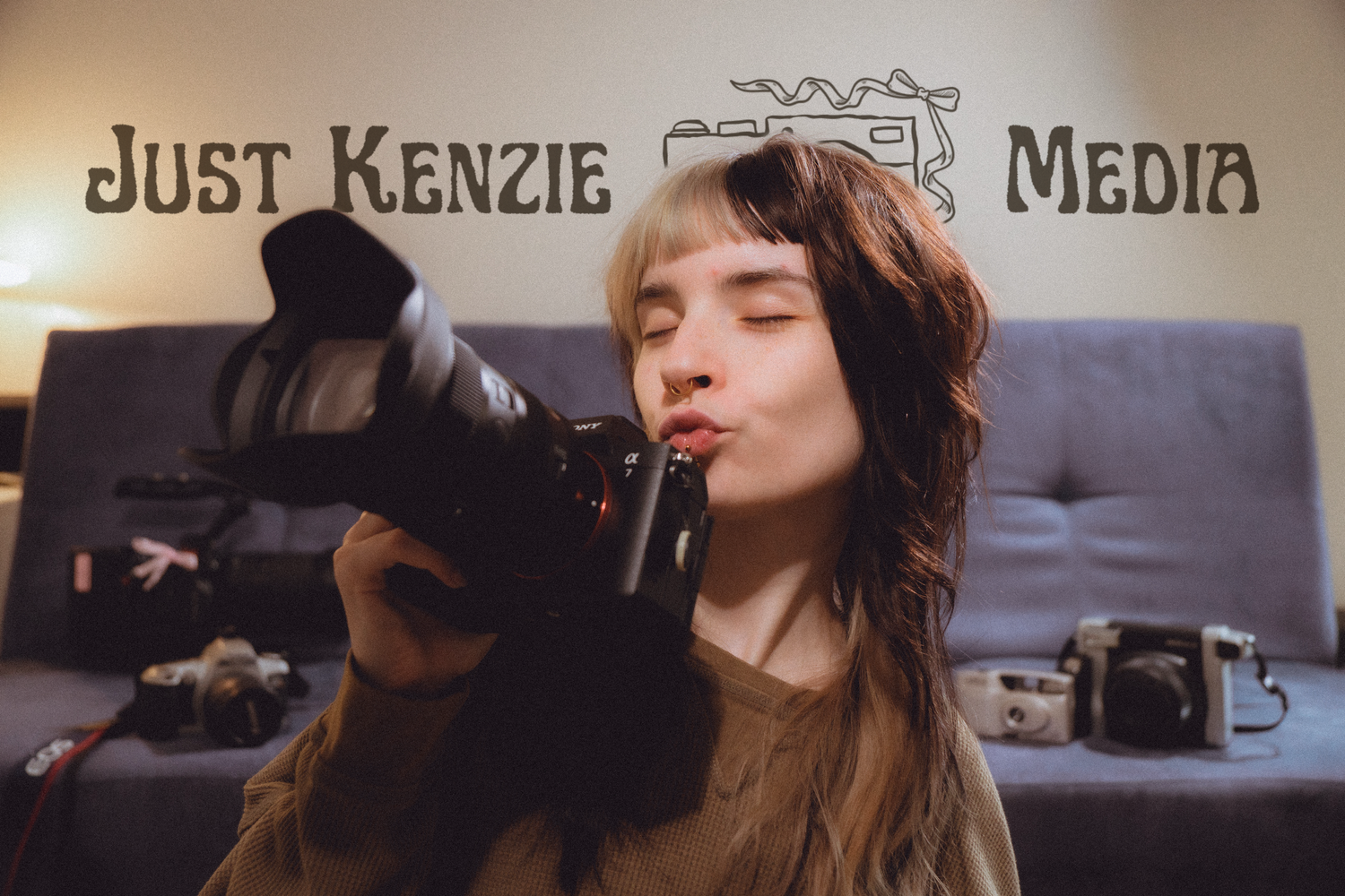 Woman kissing a professional camera in front of Just Kenzie Media logo sign indoors.