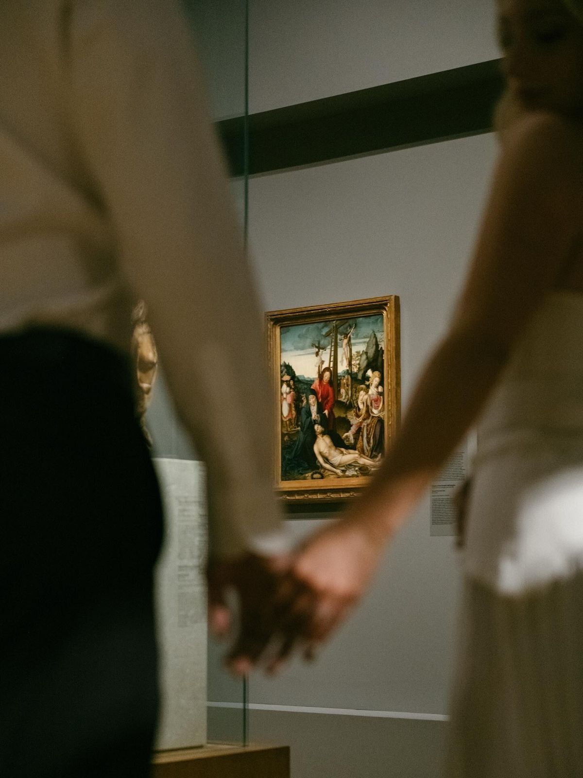 Couple holding hands in art museum gallery with classical religious painting displayed on wall in background.