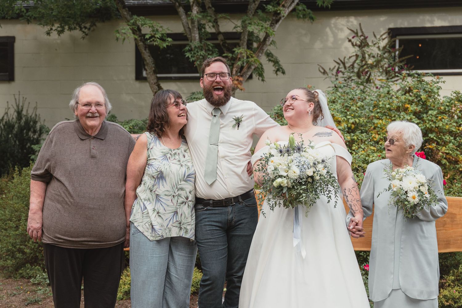 Bride and groom laugh joyfully with elderly family members outdoors at a rustic wedding celebration.