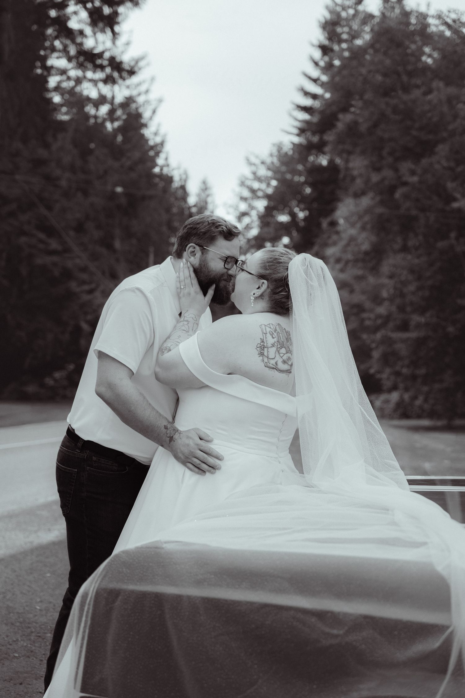 Black and white photo of a bride and groom sharing a romantic kiss on a tree-lined road, her veil flowing in the breeze.