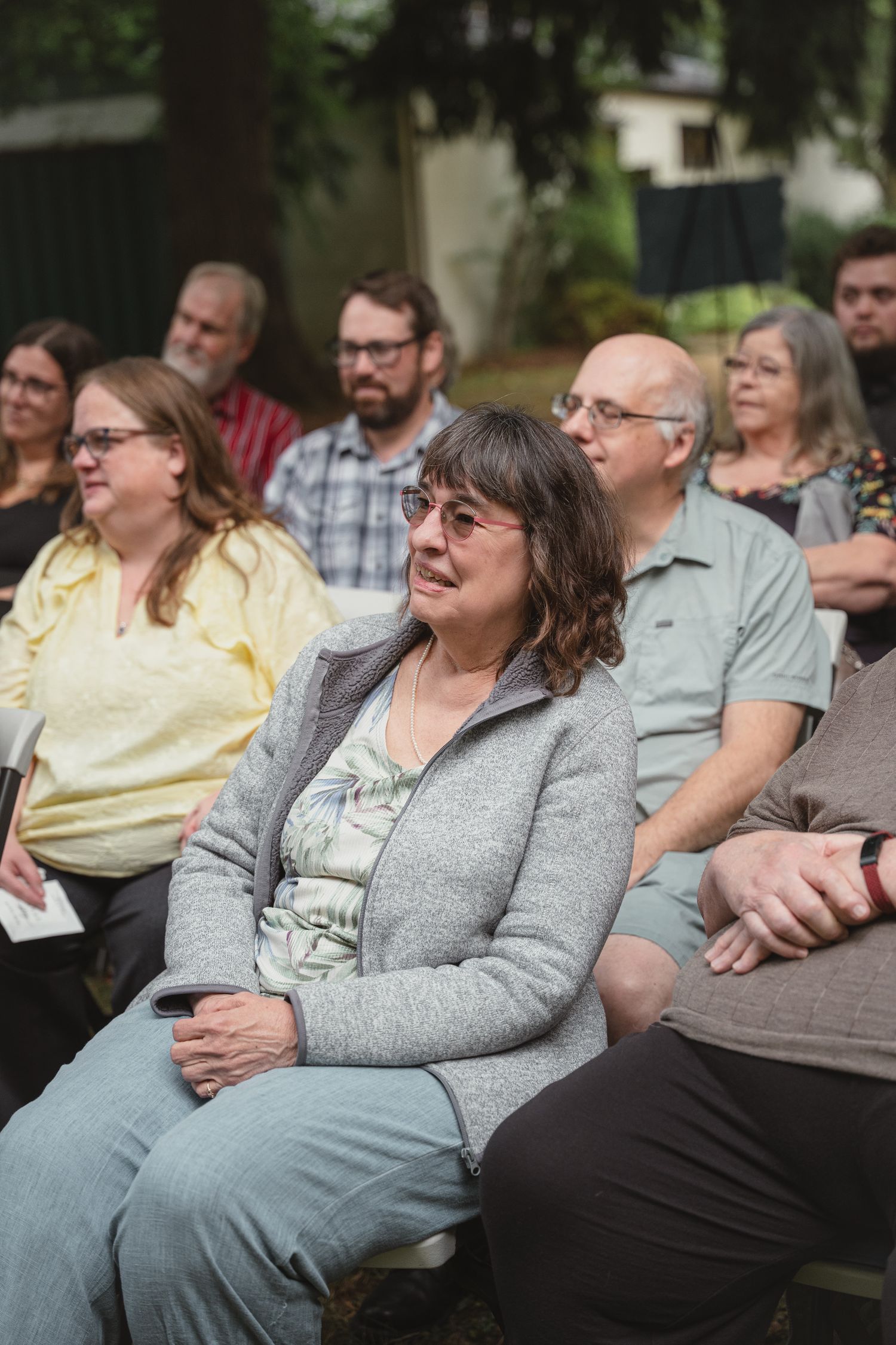 Wedding guest smiling warmly while seated at outdoor ceremony, wearing gray cardigan among other attendees.