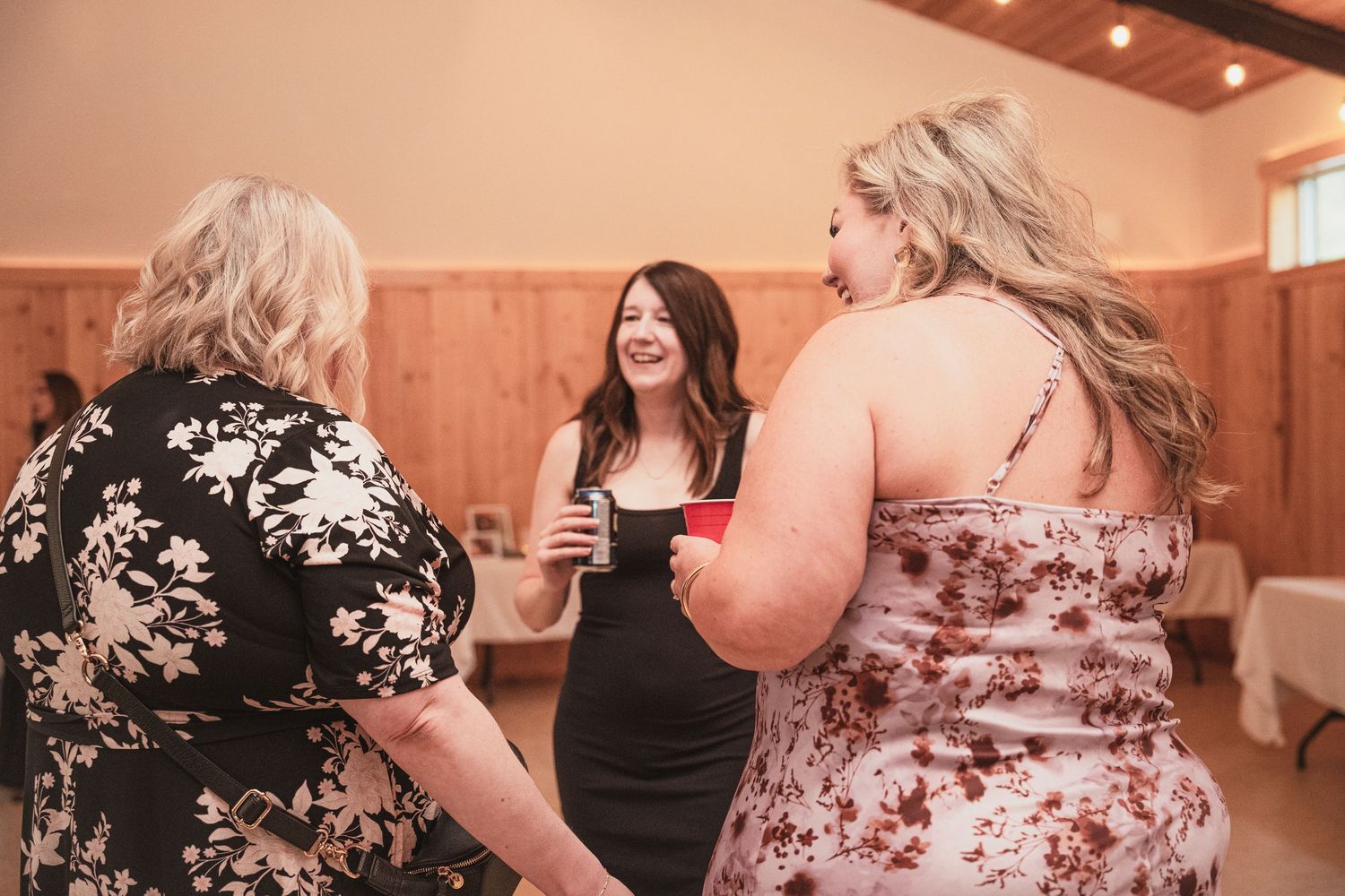Three women chatting and smiling together at wedding reception in warmly lit rustic barn venue.