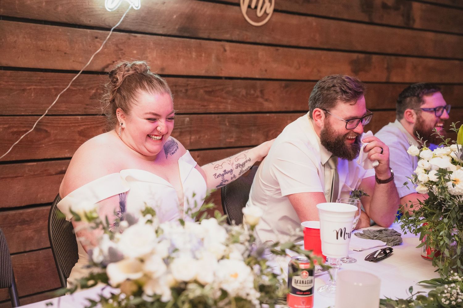 Bride and groom laugh joyfully at the head table adorned with white flowers at their wedding reception.