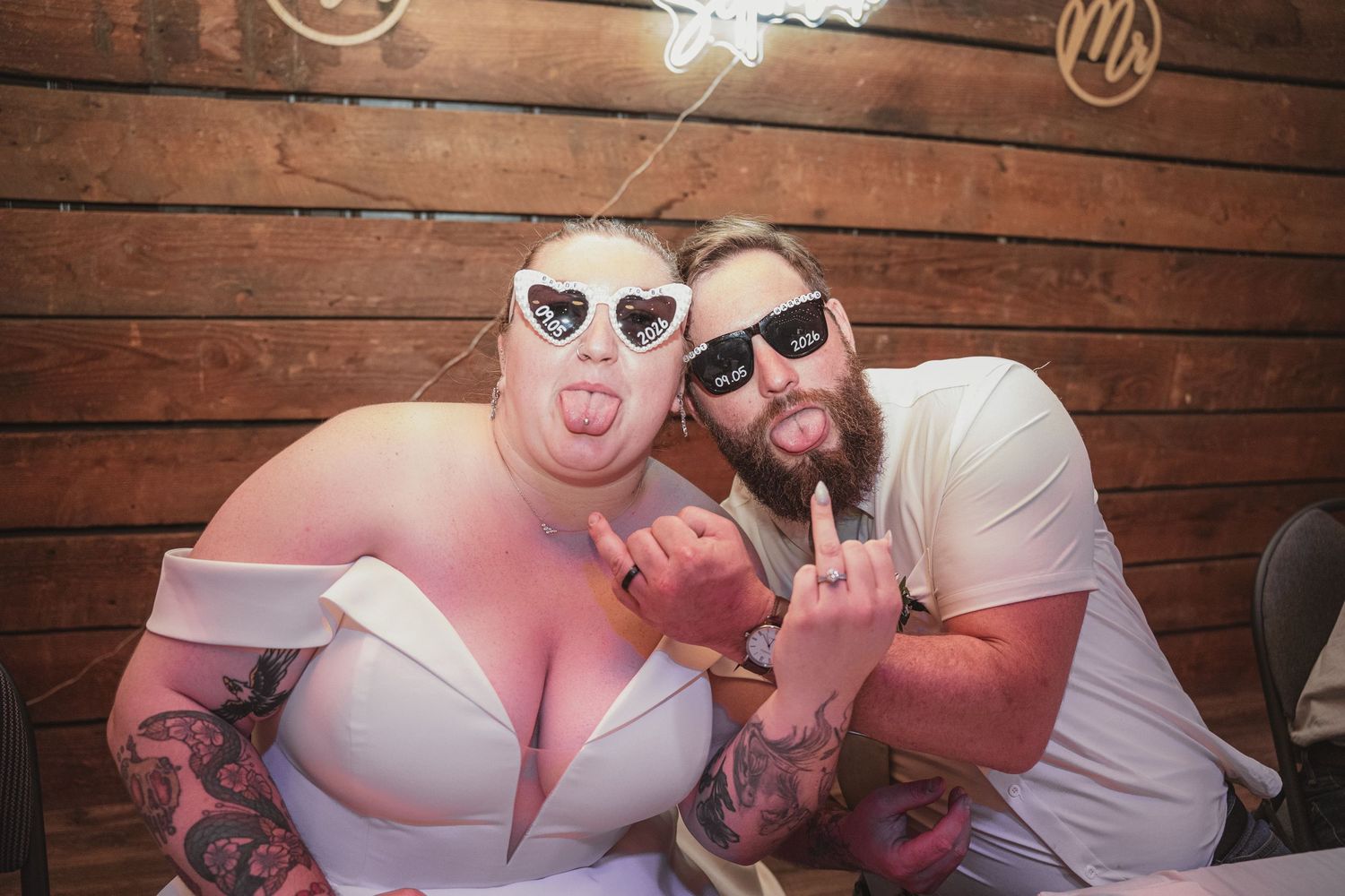Two tattooed people in heart-shaped sunglasses making rock signs at a rustic wooden venue party.