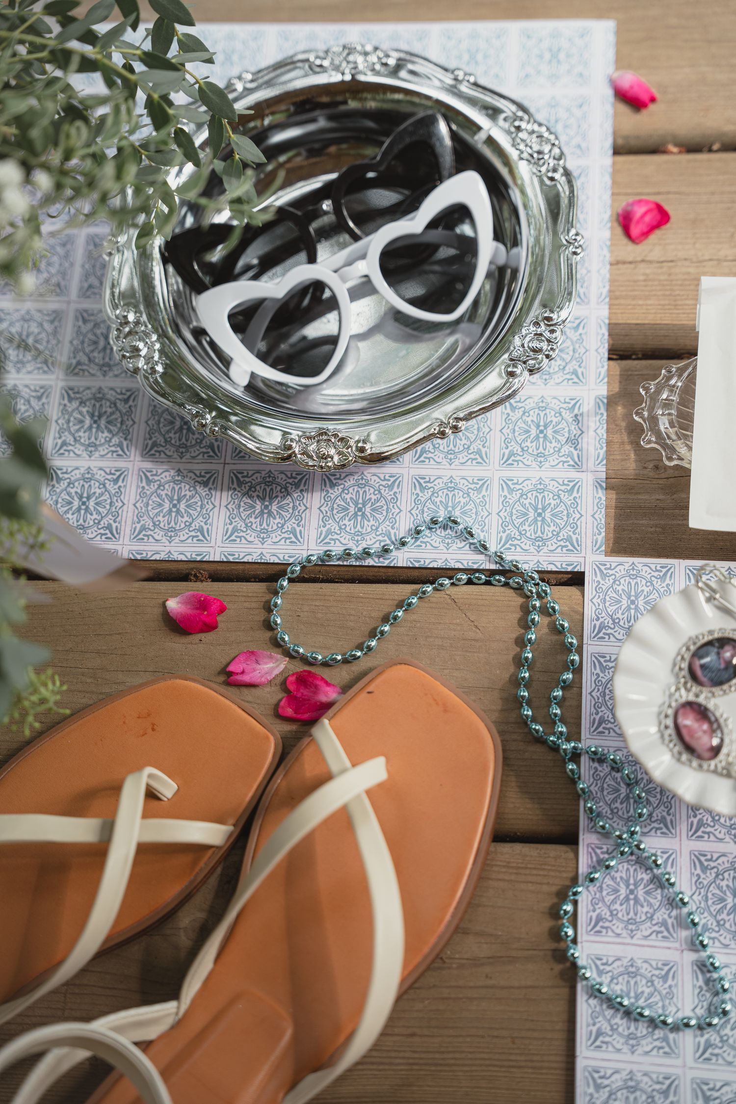 Flat lay of summer accessories including heart-shaped sunglasses, tan sandals, green beaded necklace, and rose petals on wooden surface.