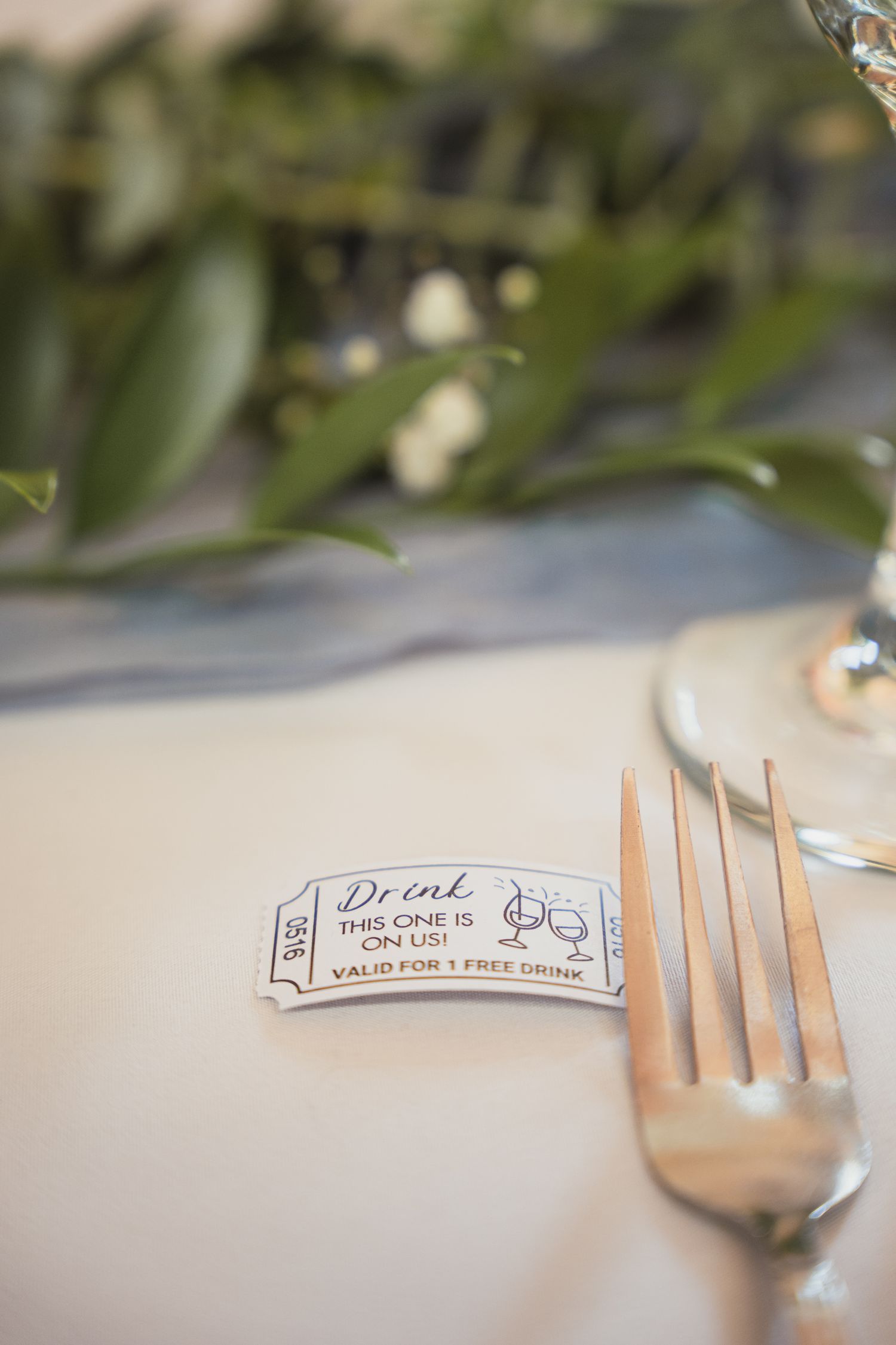 A free drink ticket placed beside a silver fork on a white linen tablecloth at an elegant event setting.
