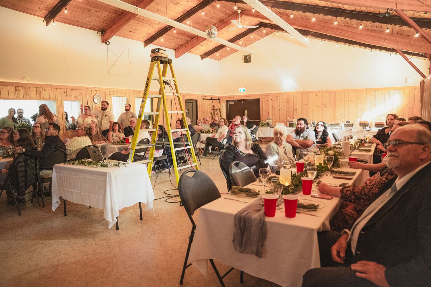 Guests seated at decorated tables in a rustic barn venue with a yellow ladder and wooden ceiling beams.