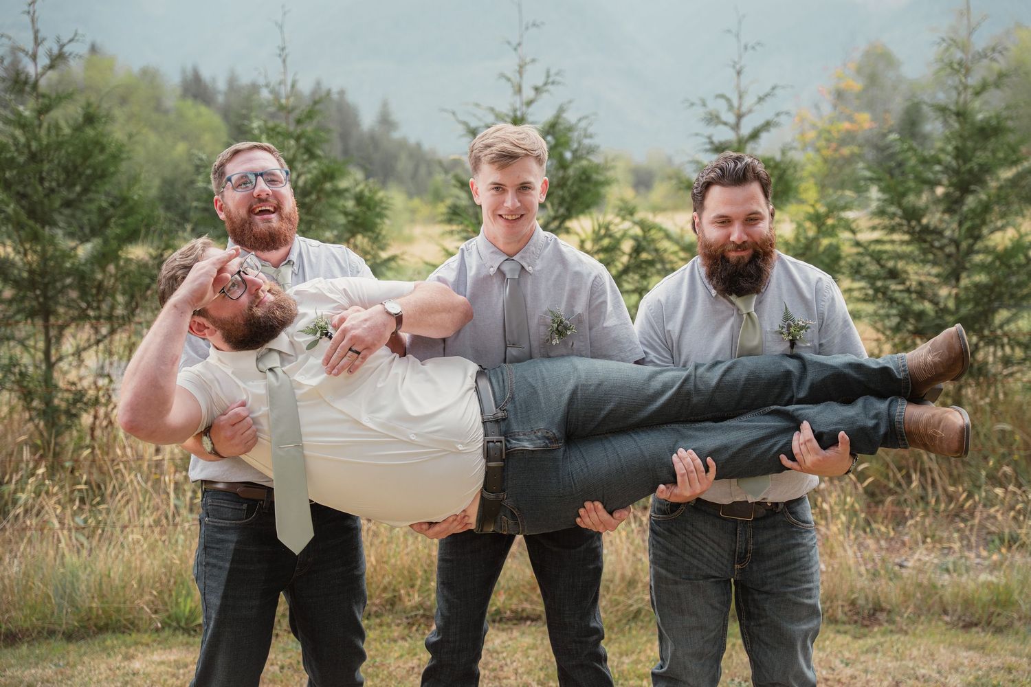 Groomsmen laughing while carrying groom horizontally at outdoor rustic wedding celebration in mountain forest setting.