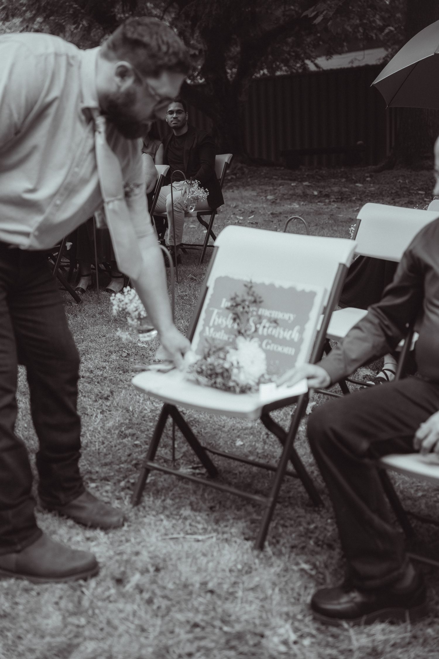 Groomsman places a floral bouquet on a memorial chair at an outdoor wedding ceremony in black and white.