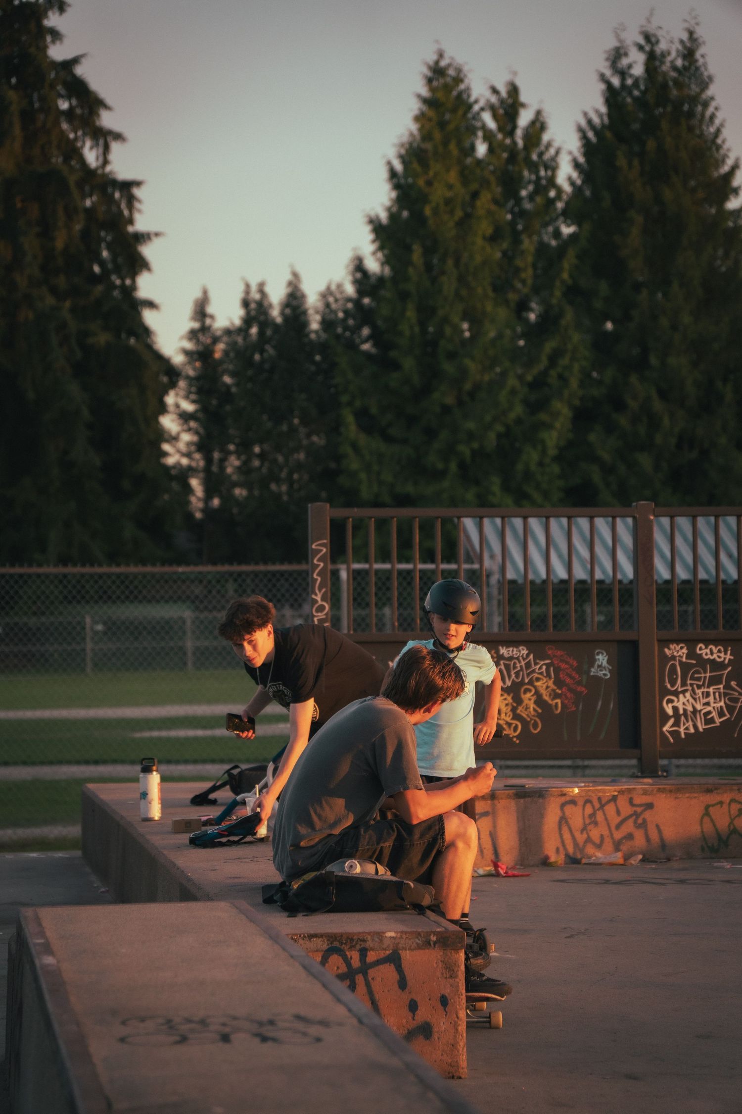 Three skateboarders resting and socializing on a ramp at an outdoor skate park during sunset.
