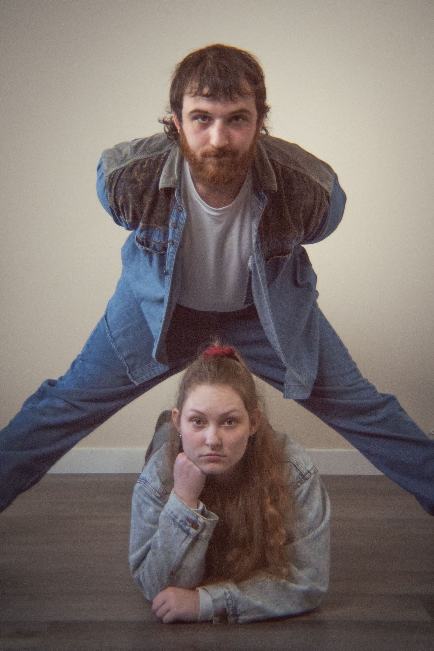 A bearded man stands over a young woman lying on the floor, both wearing denim in a quirky pose.