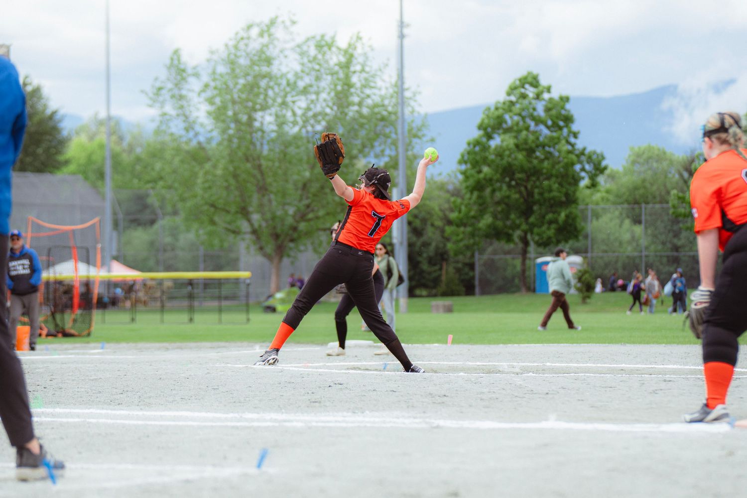 Female softball pitcher in orange uniform releases a yellow ball during an outdoor game with mountains behind.