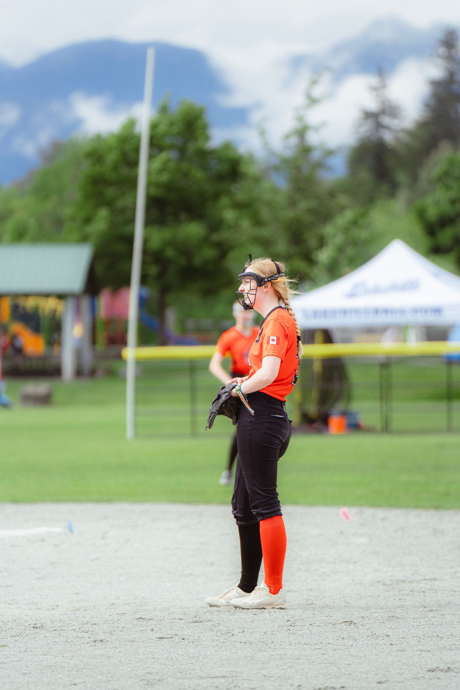 Female softball player in orange uniform and face guard standing on field smiling.