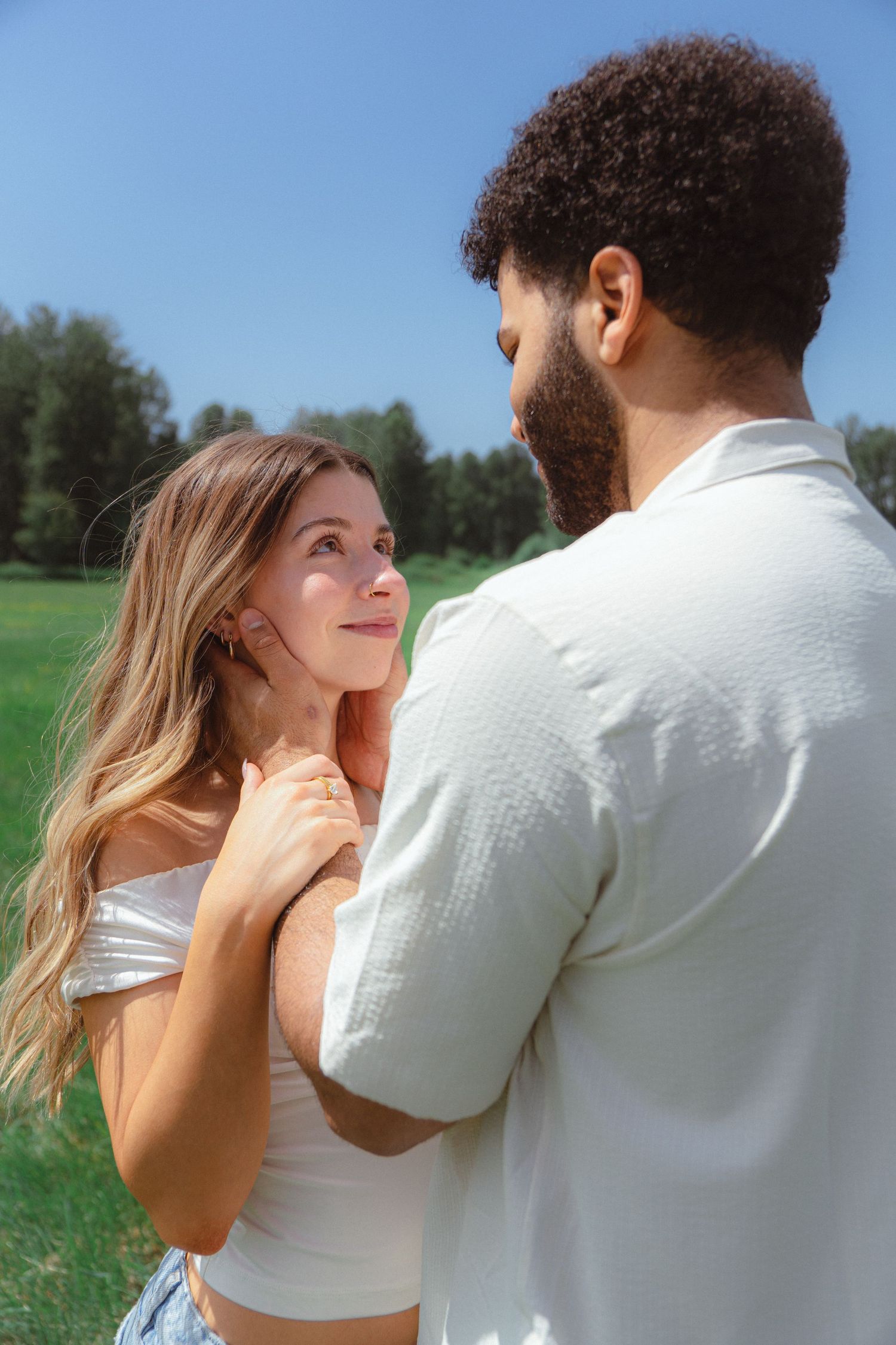 A smiling blonde woman gazes lovingly at her bearded partner while standing together in a sunny green outdoor setting.