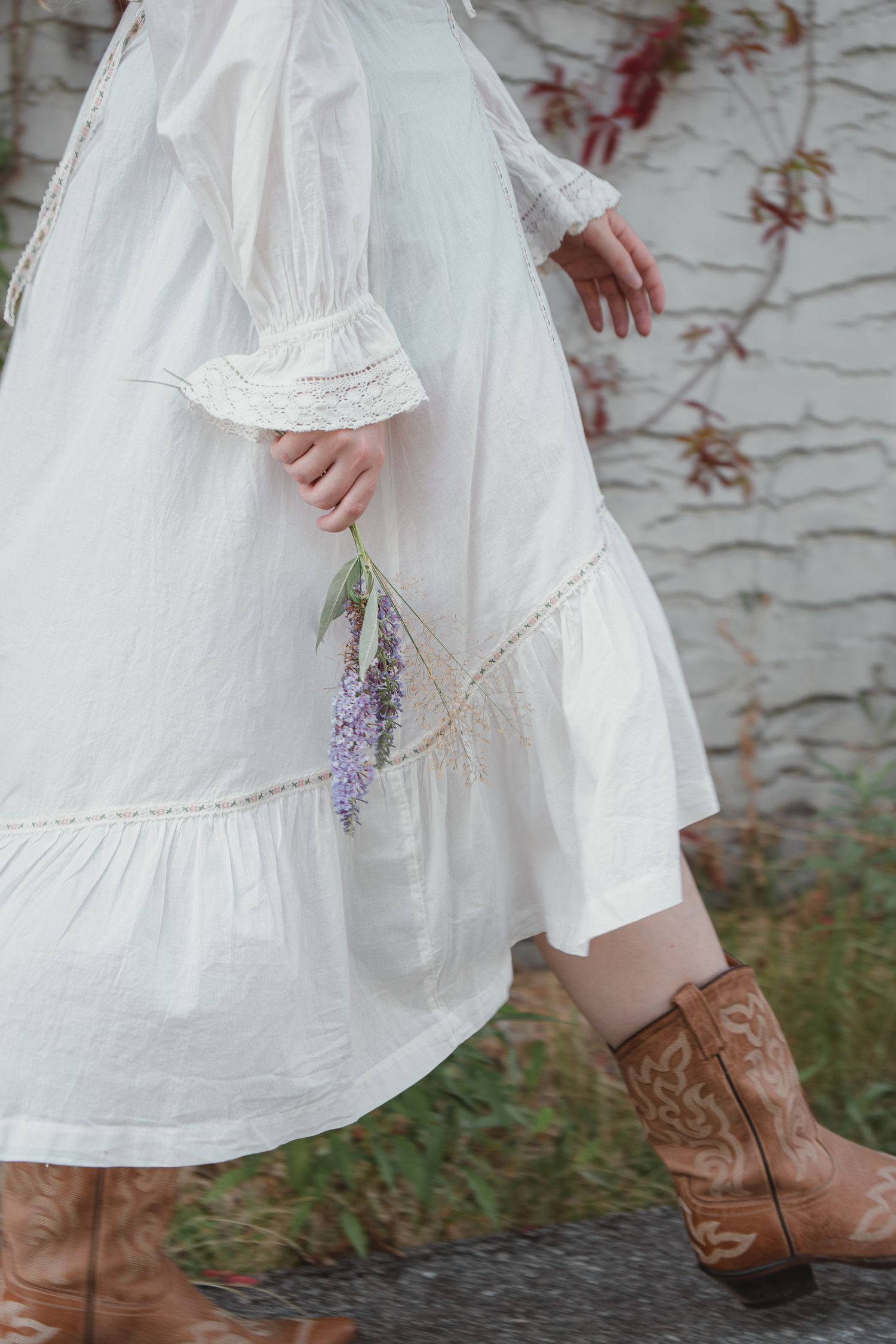 Woman in white prairie dress with lace trim and cowboy boots holding lavender flowers while walking.