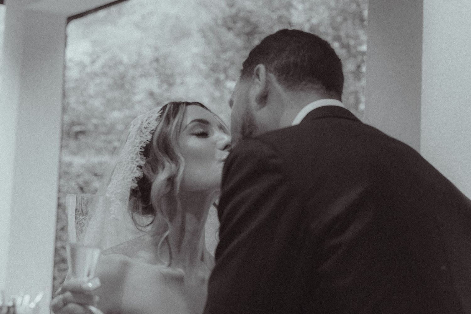 Black and white photo of bride and groom sharing a kiss while holding champagne glasses at their wedding.