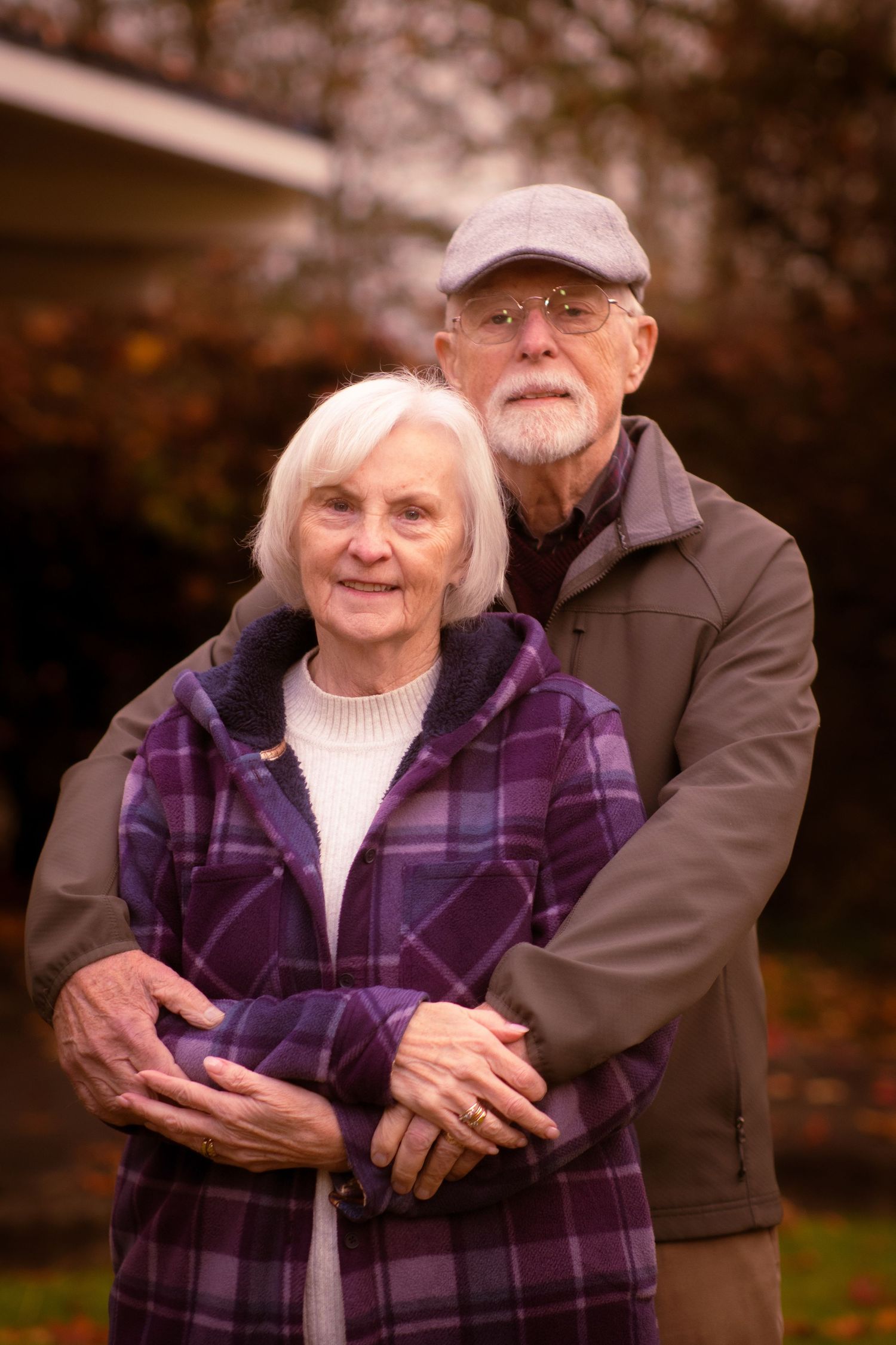 Senior couple embracing outdoors in autumn, woman in purple plaid jacket smiling warmly at camera.