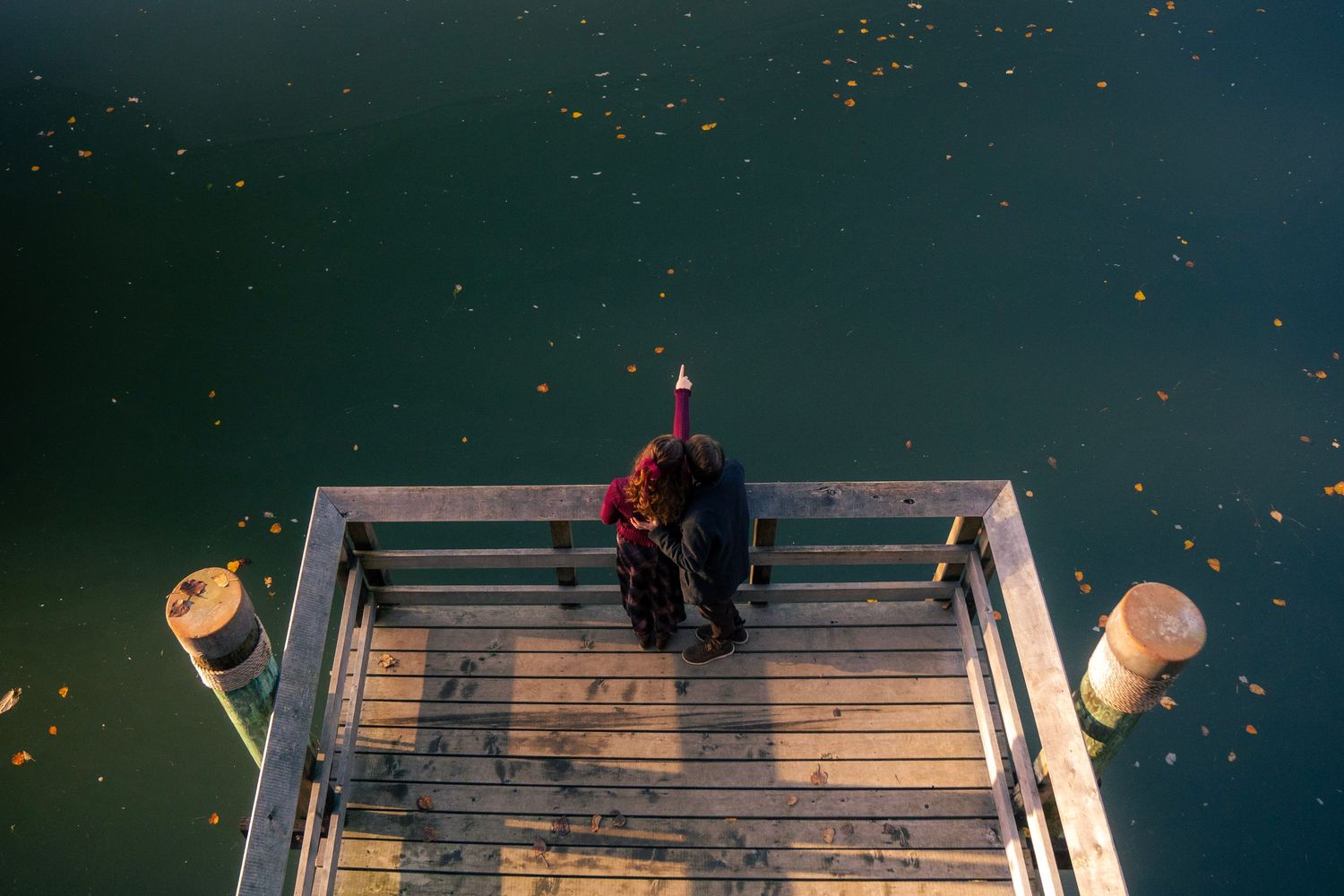 Aerial view of romantic couple standing together on wooden dock over serene green water with fallen leaves.
