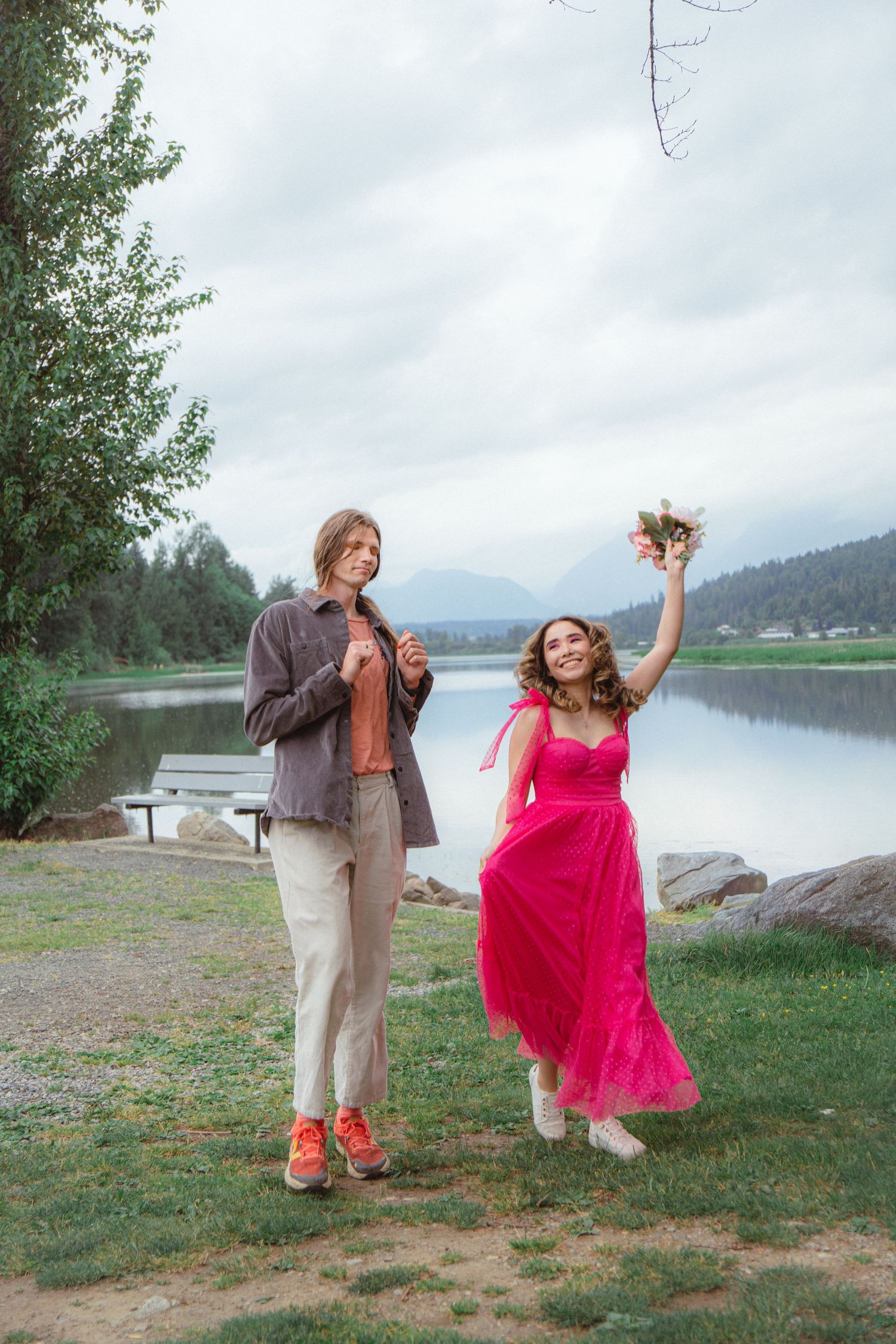 Woman in bright pink dress raising bouquet joyfully beside companion near calm reflective lake.