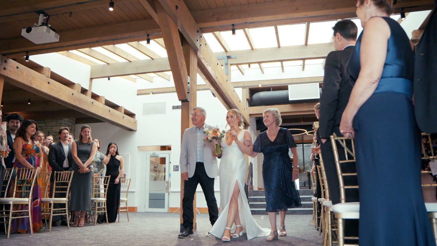 Bride walks down the aisle escorted by two family members at an indoor rustic wedding ceremony.