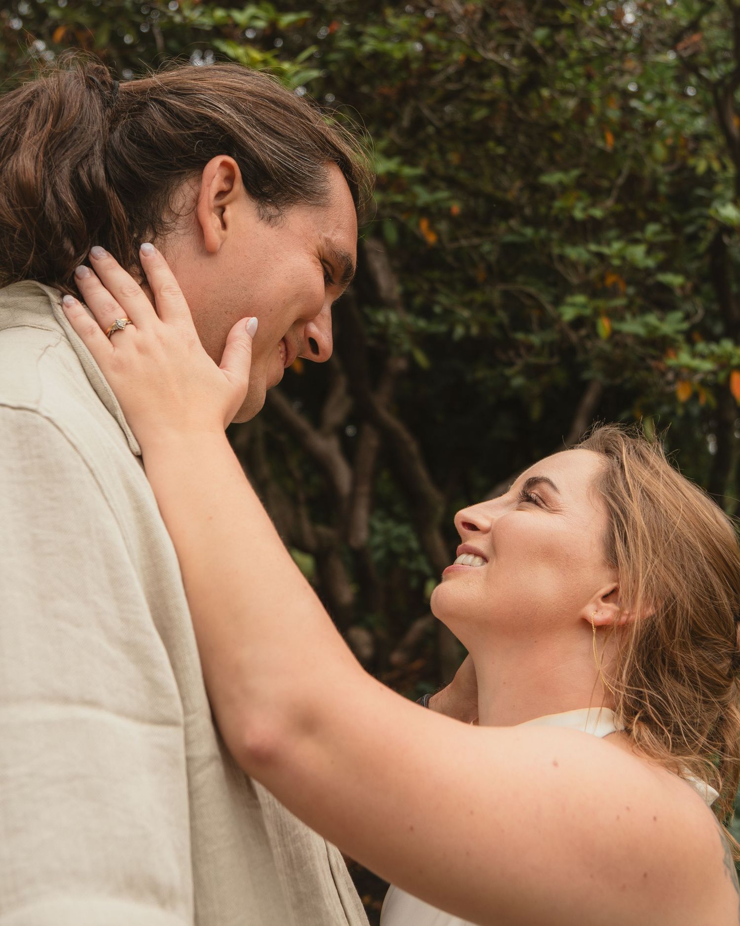 A couple shares a romantic moment outdoors, with the woman smiling up at the man while gently holding his face.