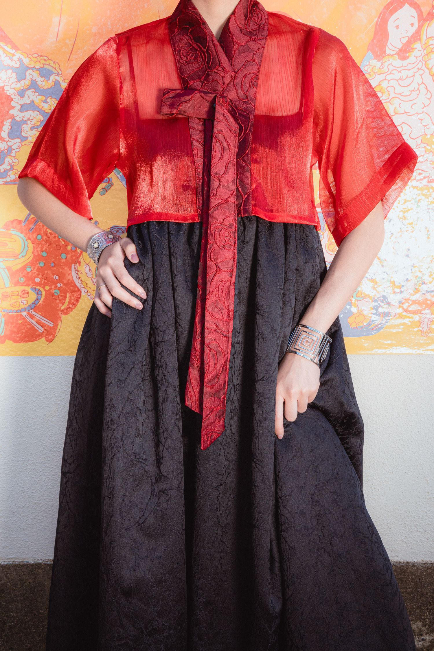 Woman wearing red sheer top with bow tie over black maxi skirt, silver bracelets, traditional Asian-inspired outfit.