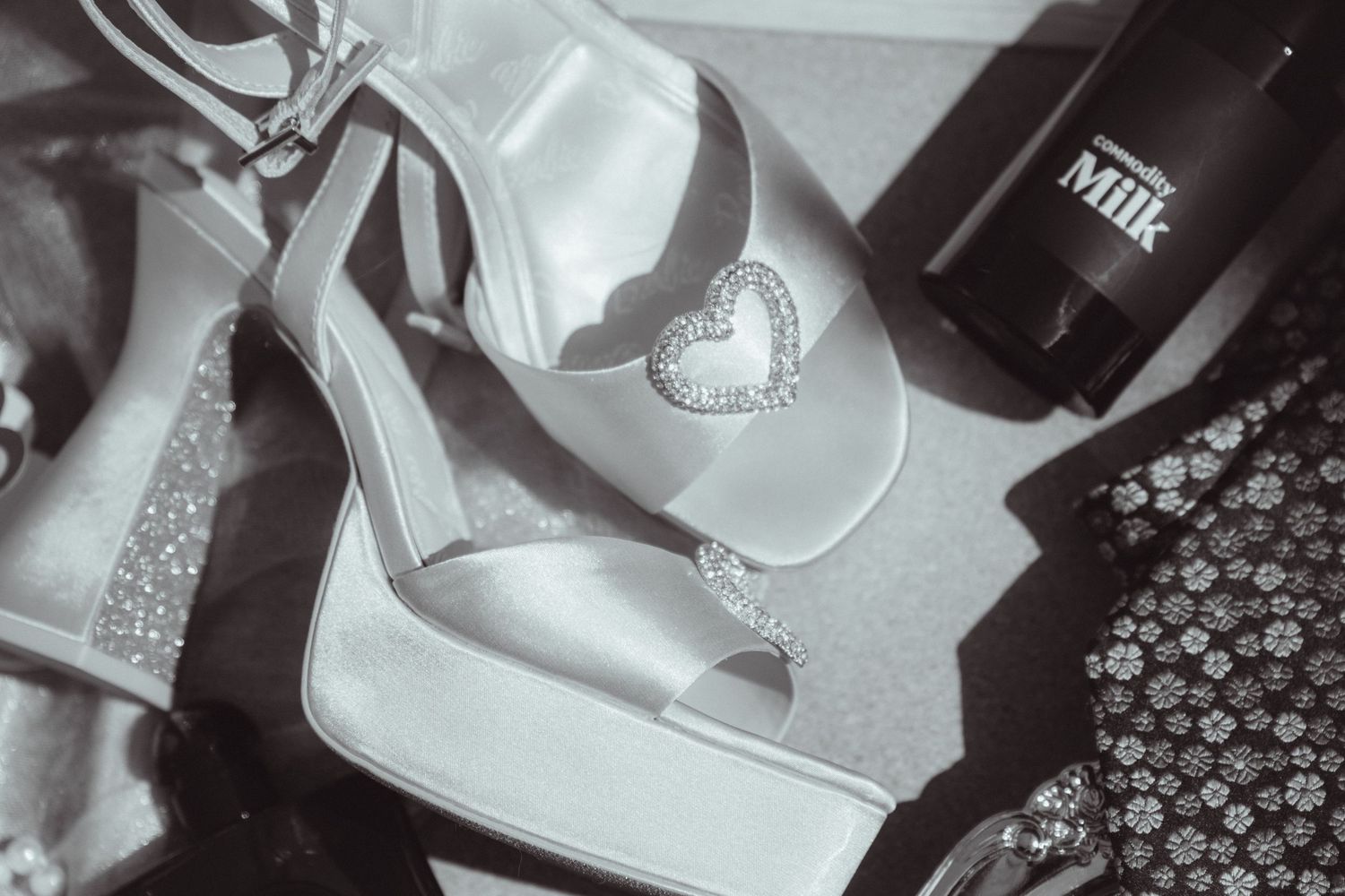 Black and white photo of elegant satin platform heels with crystal heart detail beside a Milk bottle.