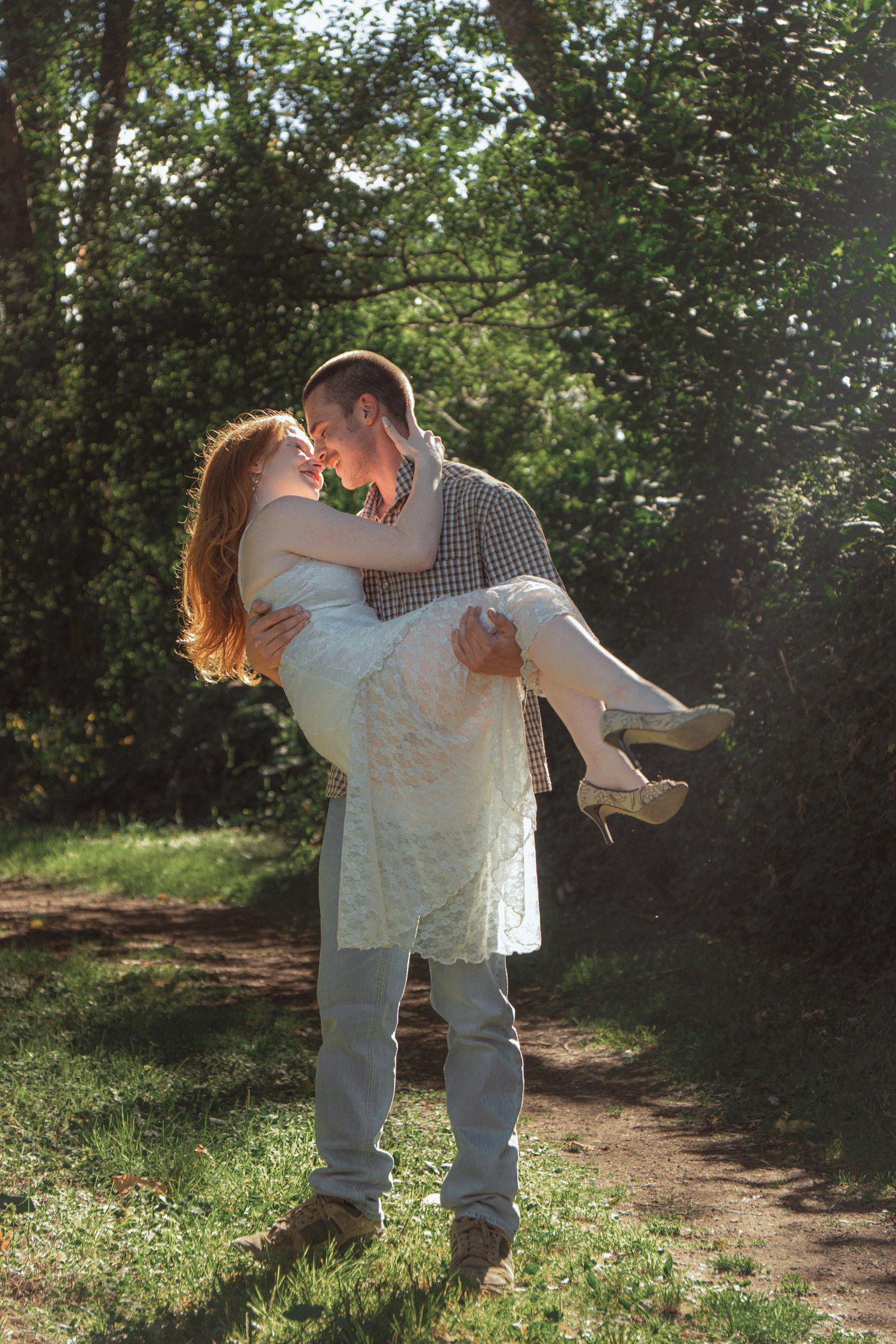 Man carrying woman in white lace dress in his arms, sharing a romantic moment in a sunlit forest.