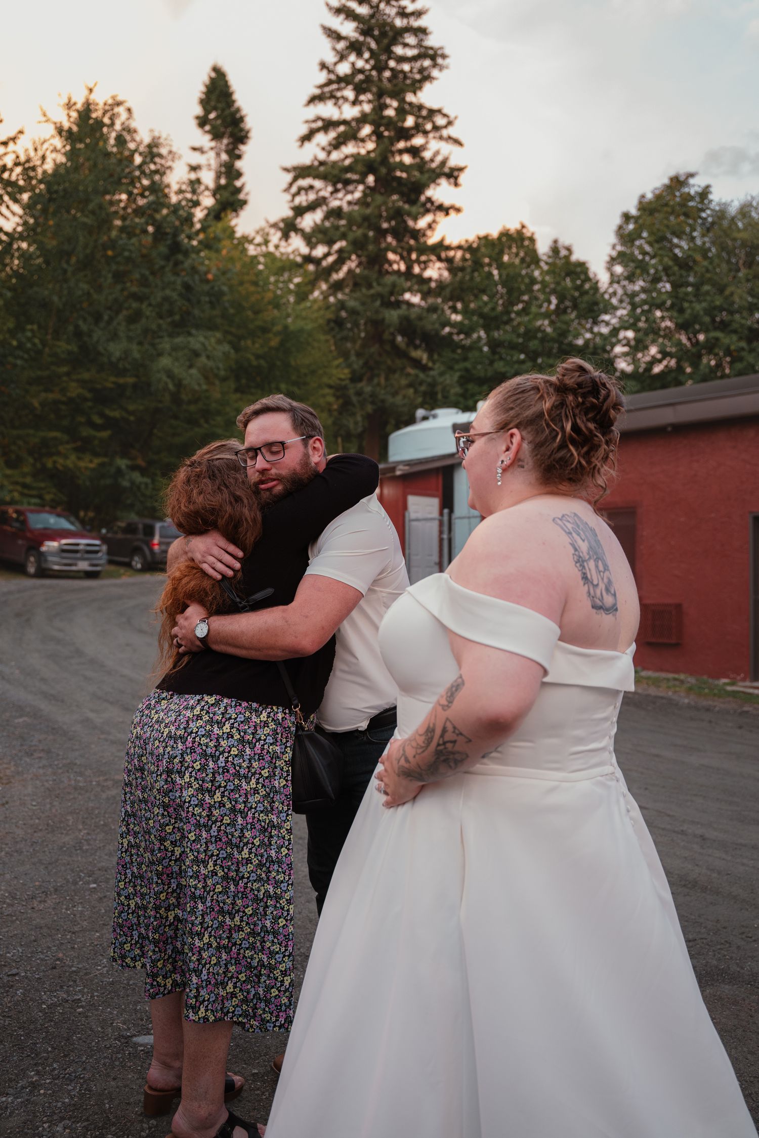 Bride in white gown watches as groom embraces a red-haired guest in an outdoor farewell at sunset.