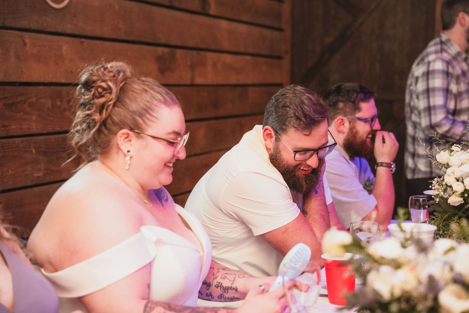 Bride and tattooed groom laugh together while seated at the wedding reception dinner table.
