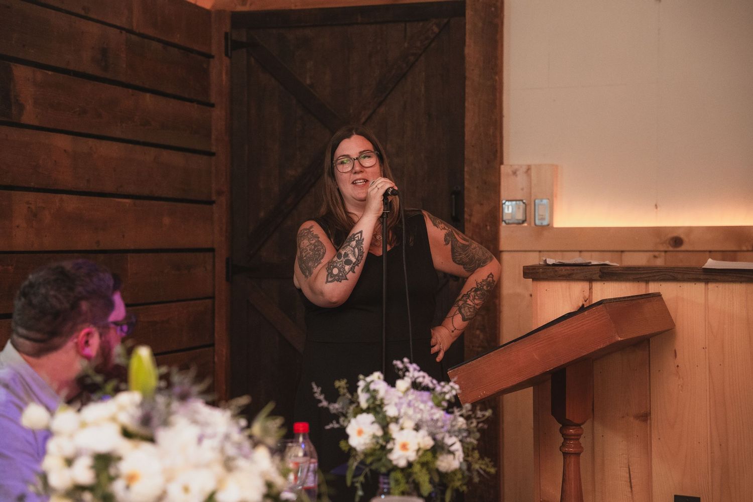 Tattooed woman speaking at a podium during a wedding reception in a rustic barn venue.