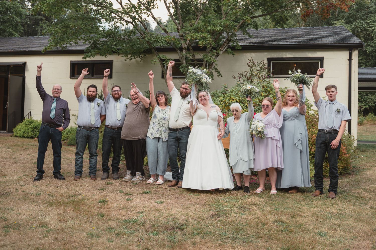 Wedding party celebrating outdoors, raising arms joyfully in front of a building, with bride in white gown and guests in coordinated attire.