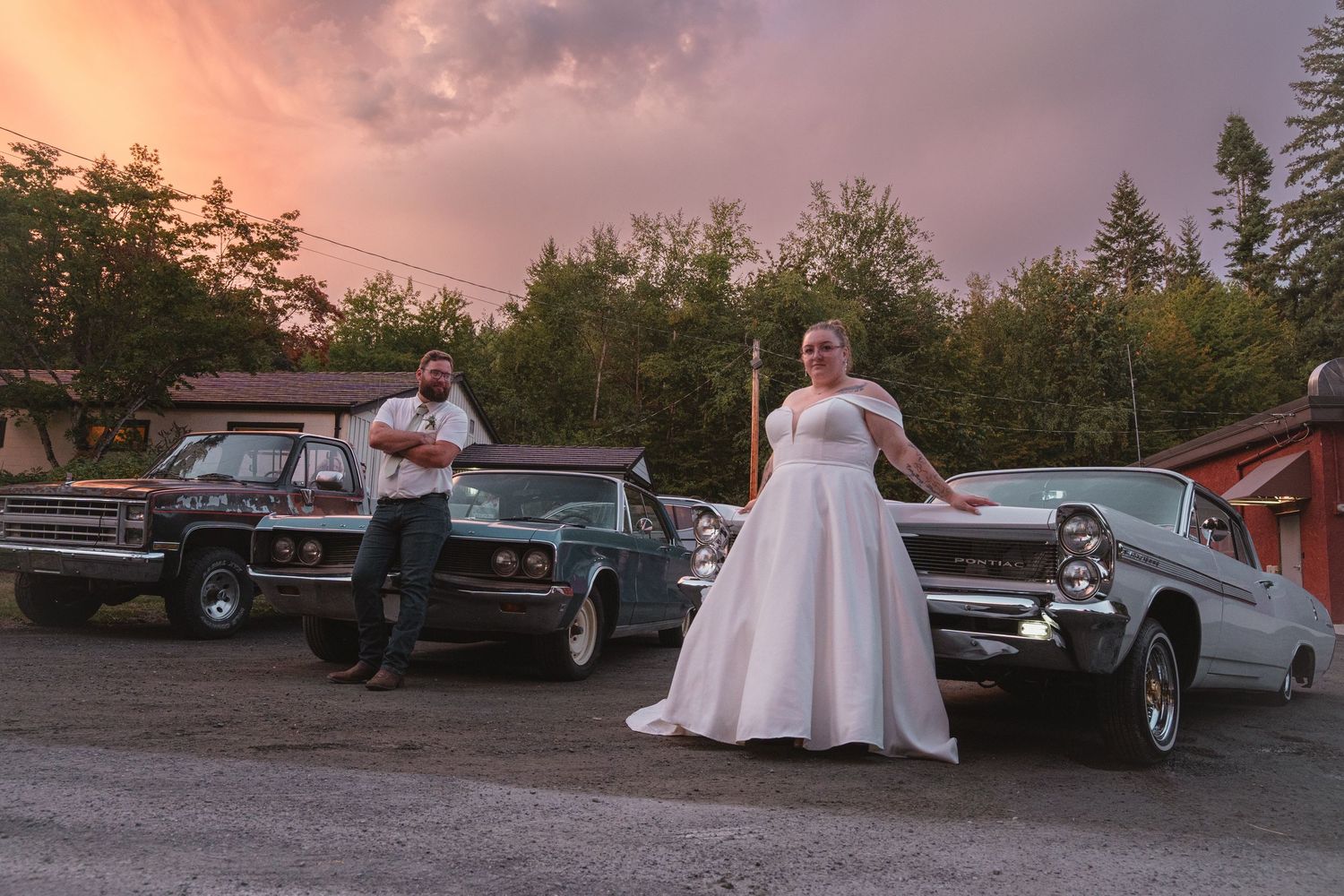 Bride in white gown and groom pose with classic vintage cars at sunset in a scenic outdoor parking area.